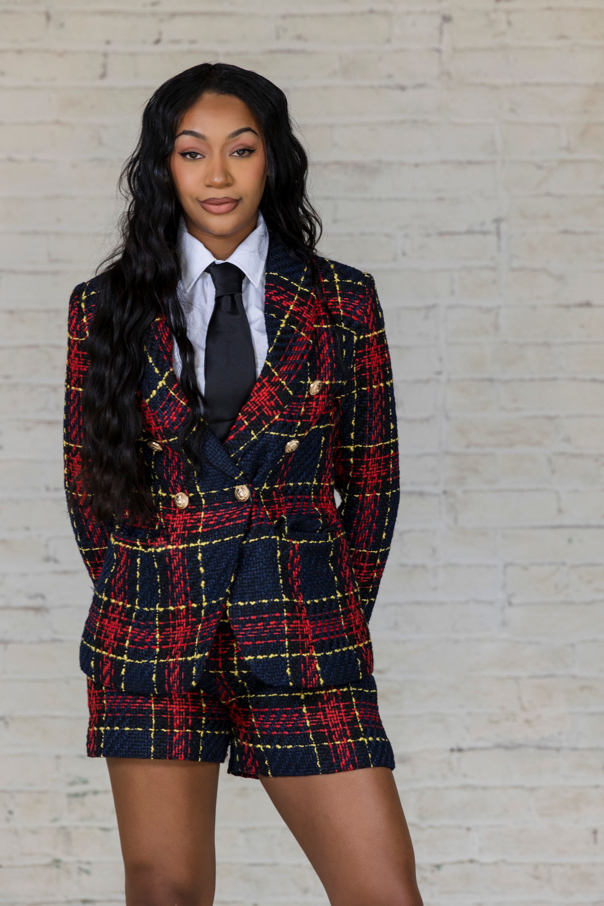 Person standing against a white brick wall, wearing a red-and-black plaid blazer and skirt with a white shirt and black tie.