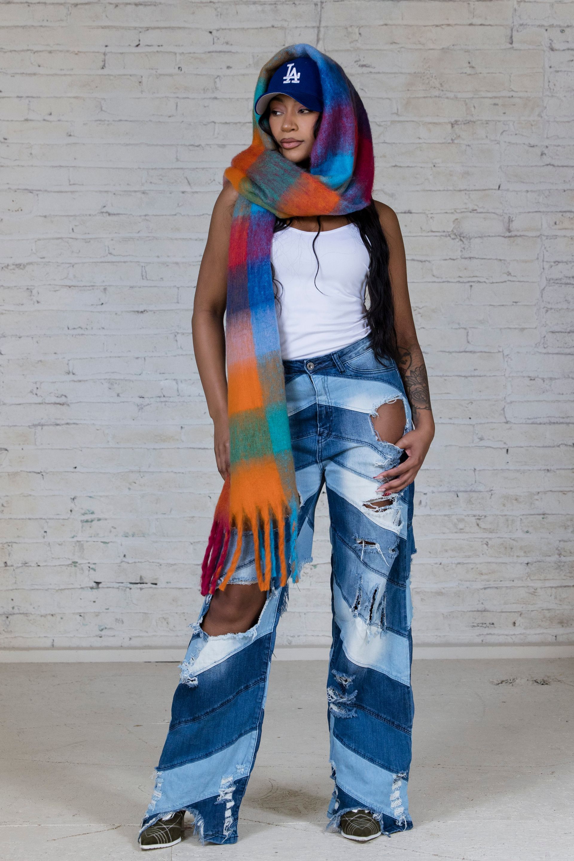Person in colorful scarf, white top, and blue patchwork jeans standing against a brick wall.