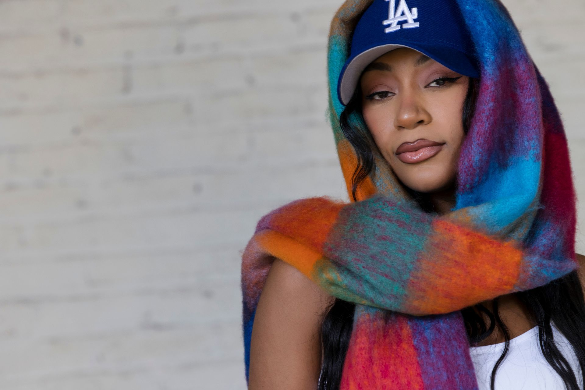 Person in a blue LA cap and multicolored scarf against a light brick wall.