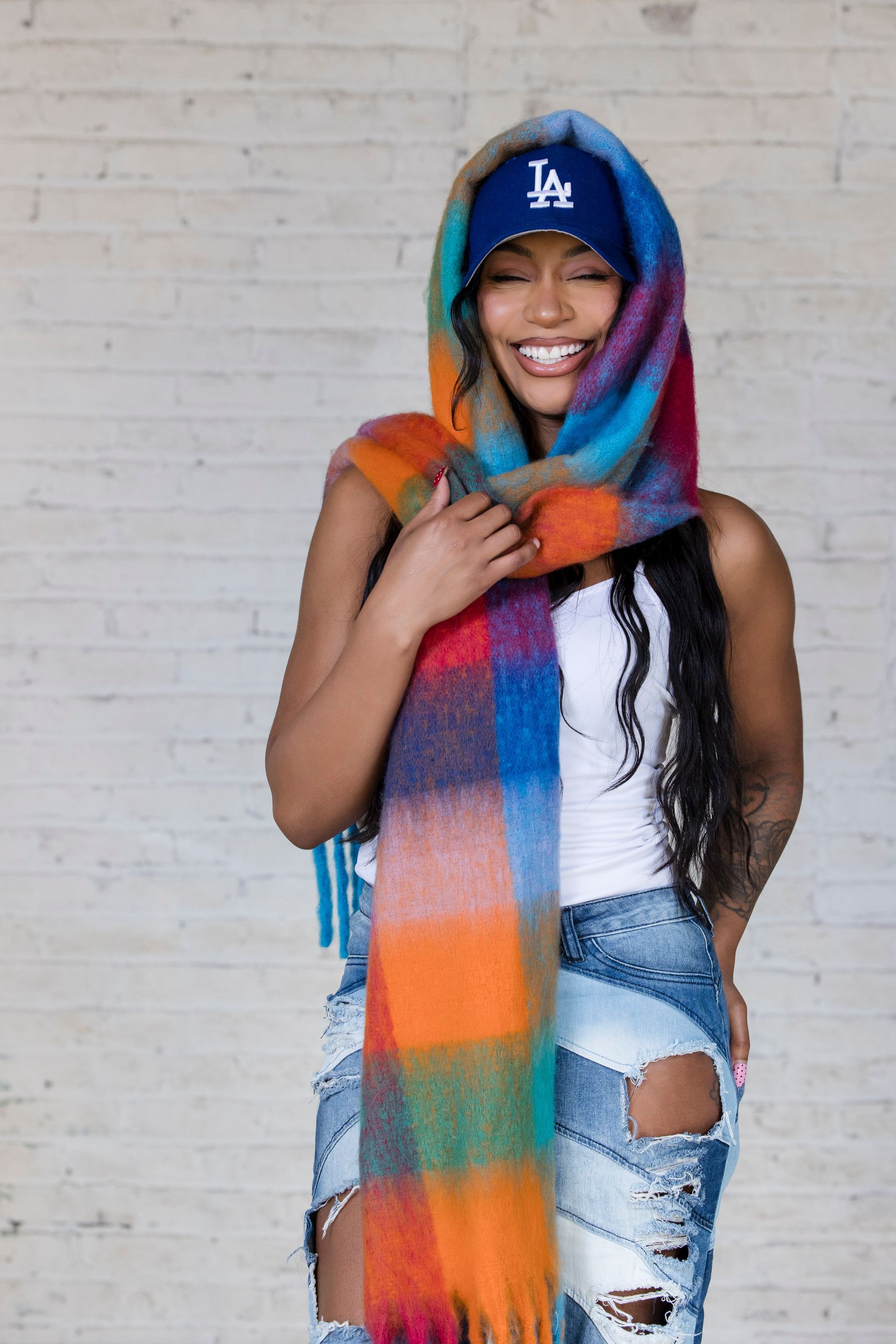 Woman in a blue cap and rainbow scarf, smiling against a white brick wall.