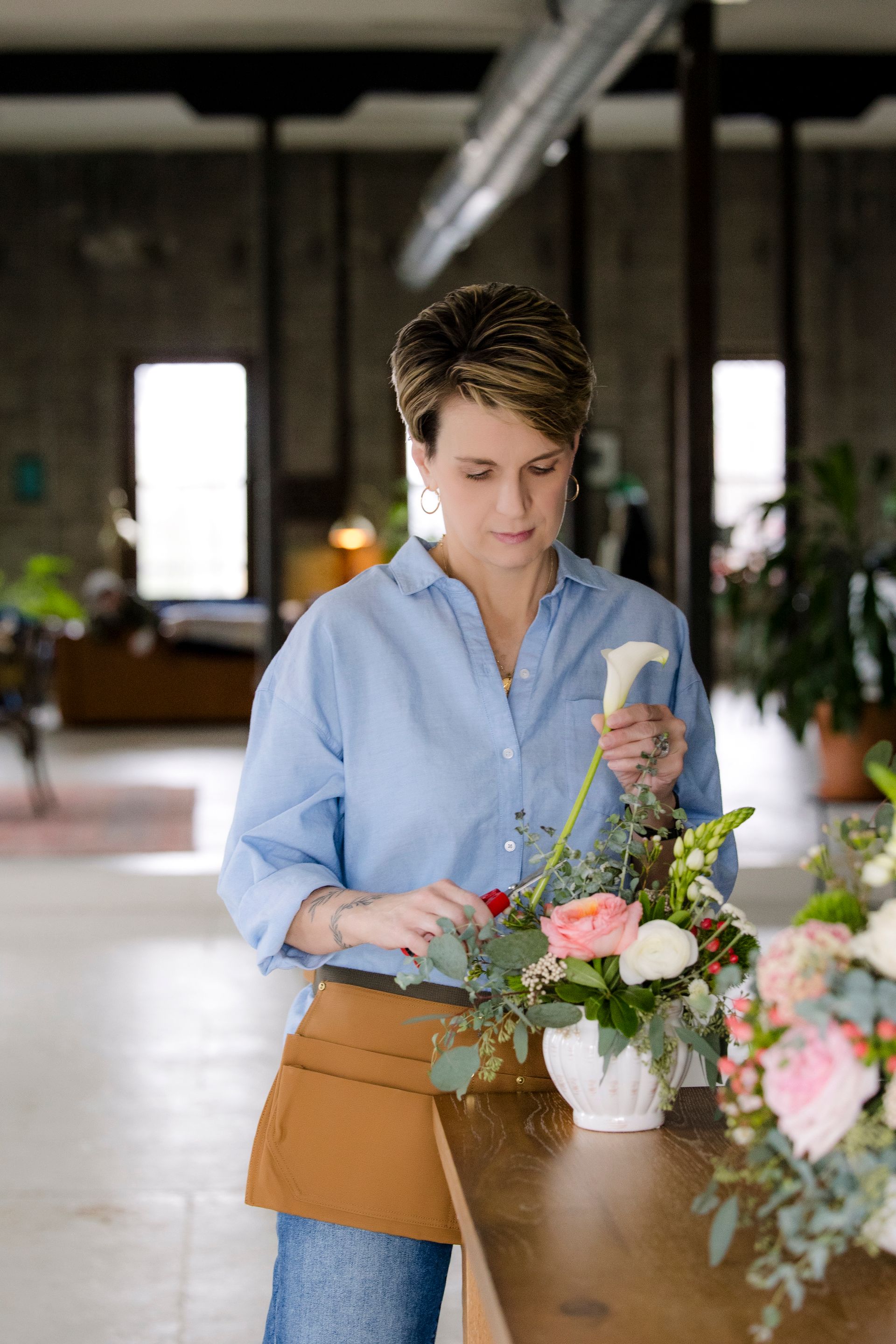 Person arranging flowers at a wooden table in a bright workshop, wearing a blue shirt and apron