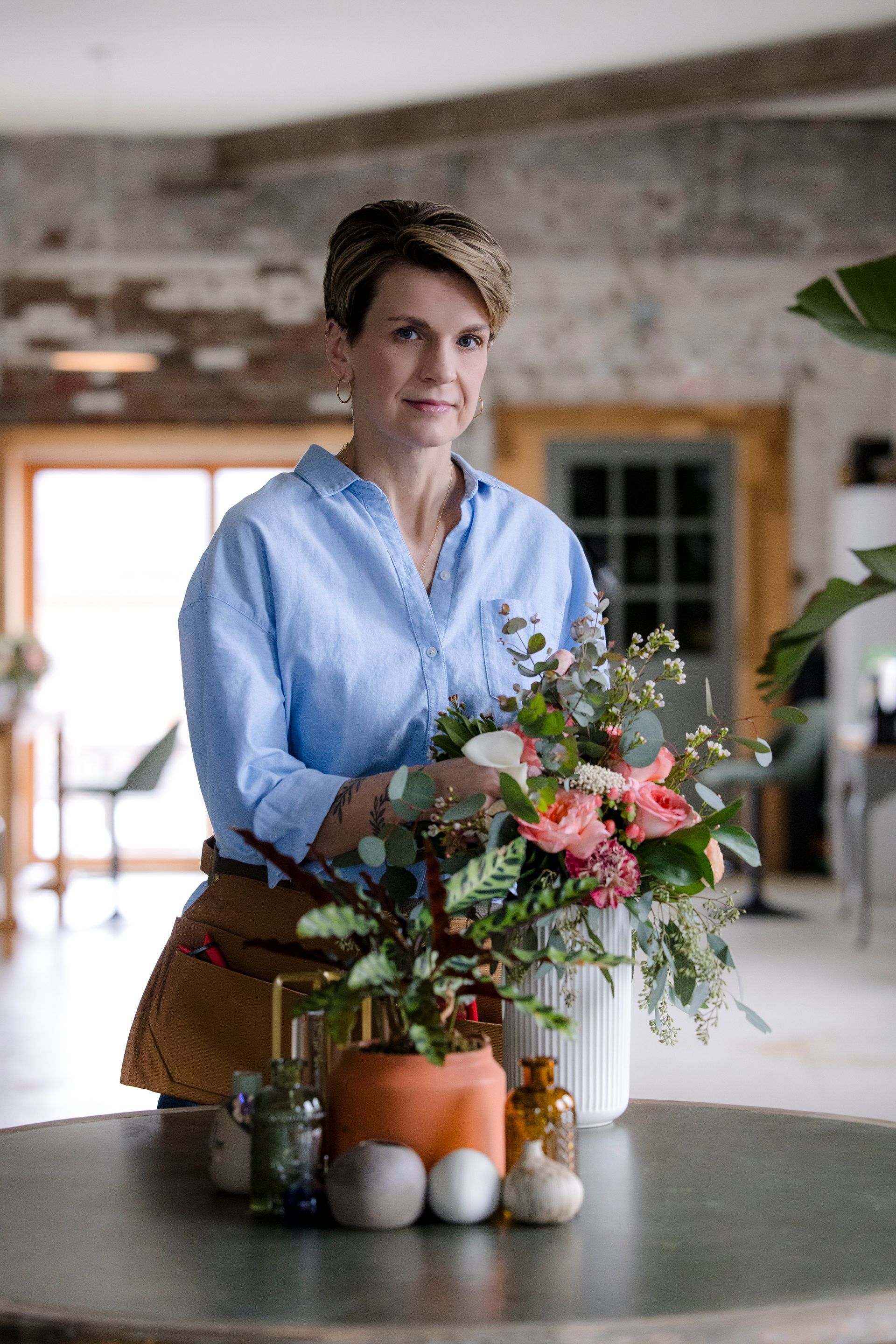 Person in a light blue shirt arranging flowers on a table with pumpkins in a rustic room