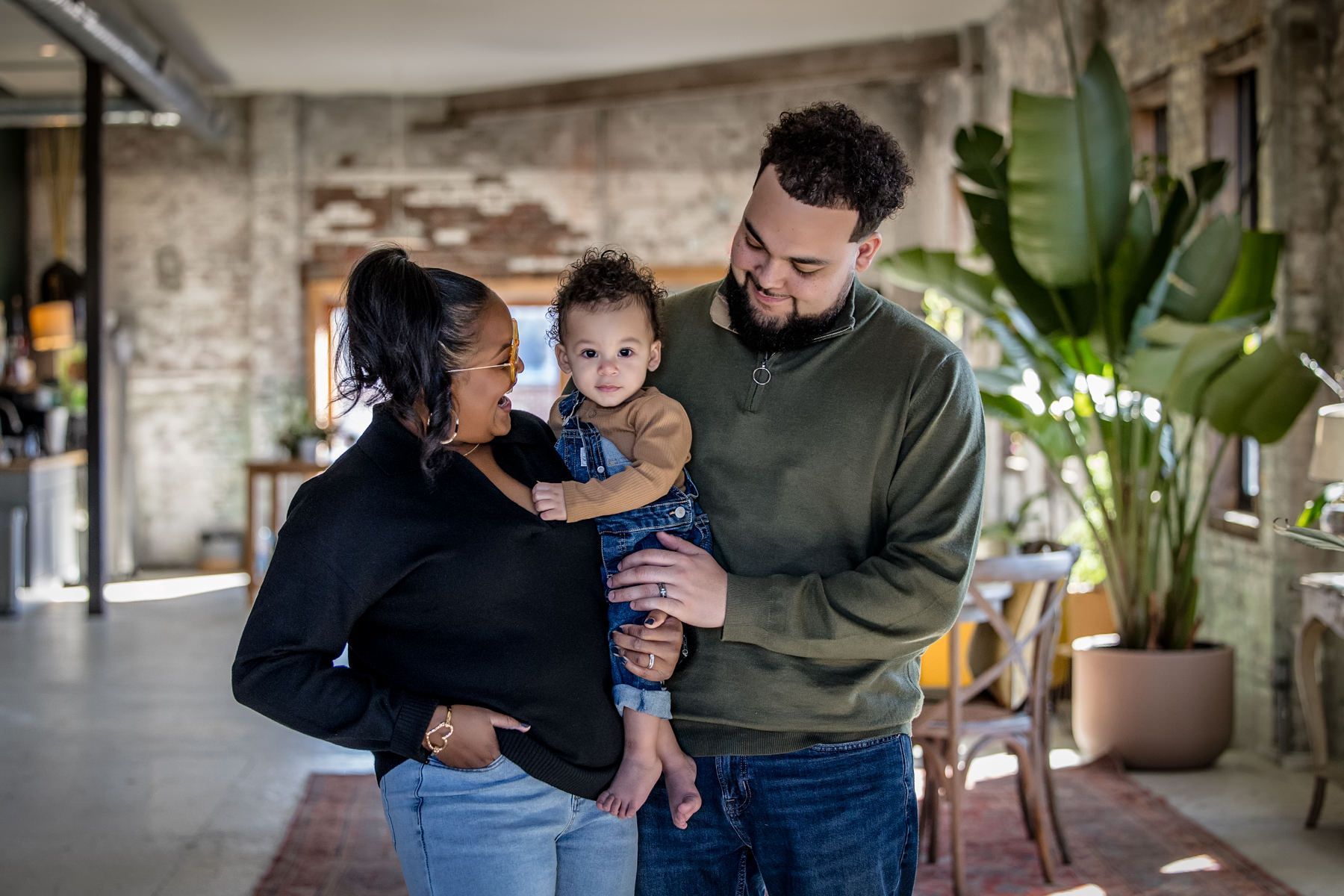 Family of three standing indoors, smiling and holding a small child near large potted plants.