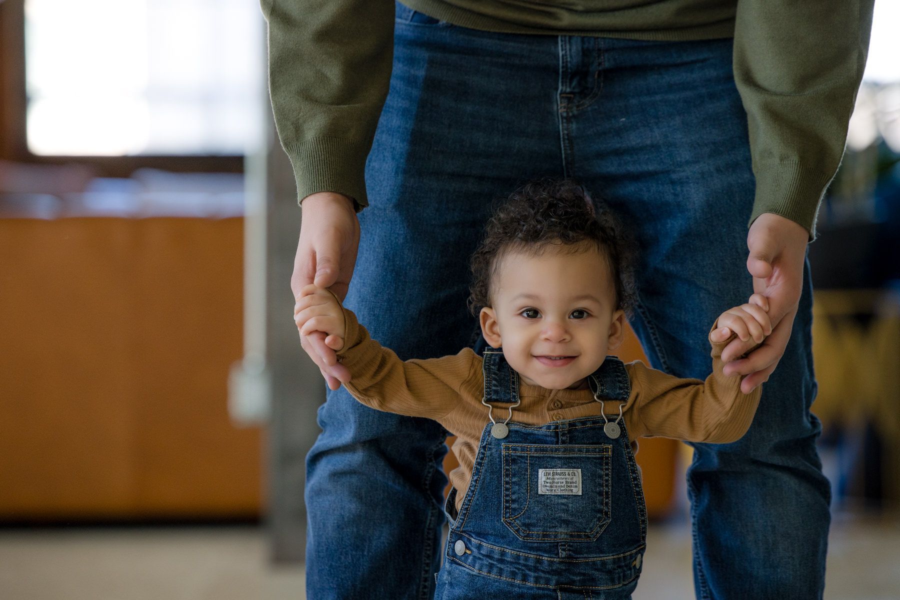 Adult holding a smiling toddler's hands as they walk indoors, with jeans and overalls visible.