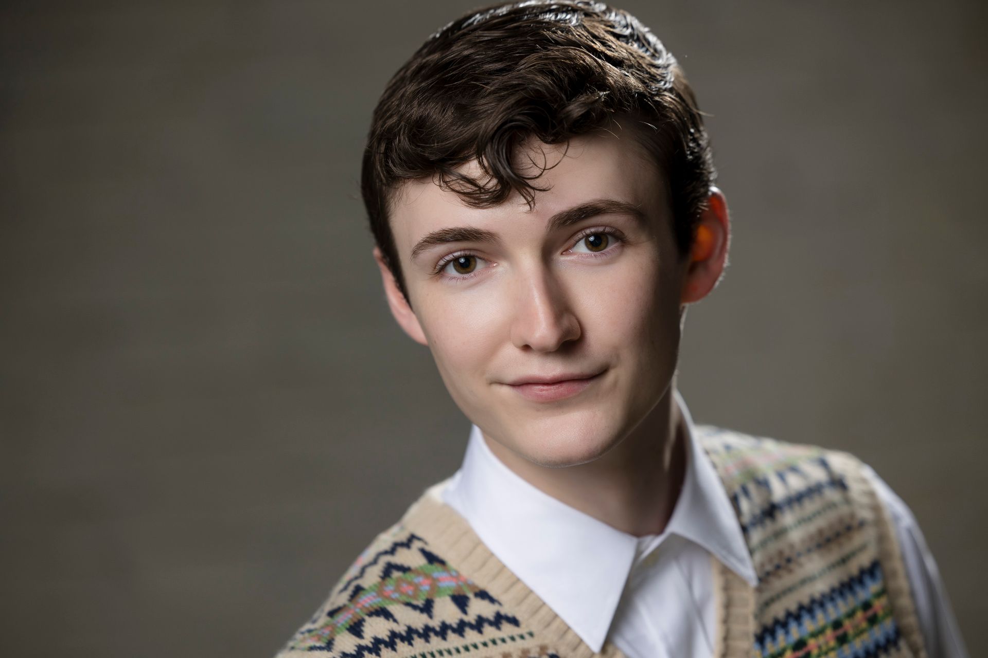 Portrait of a person with short dark hair wearing a patterned sweater over a white collared shirt, smiling softly.
