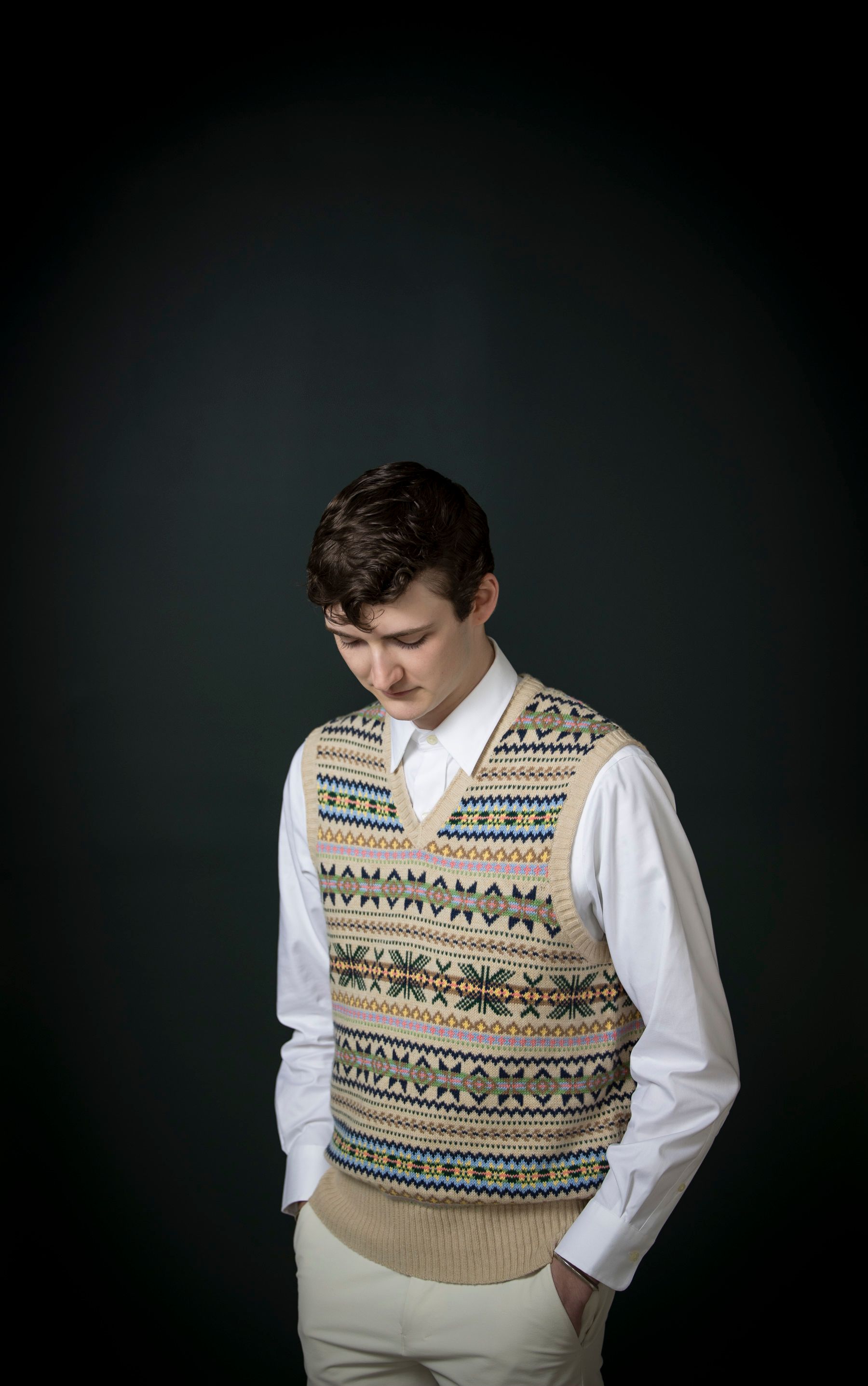 Man in a patterned sweater vest and white shirt looking down against a dark background