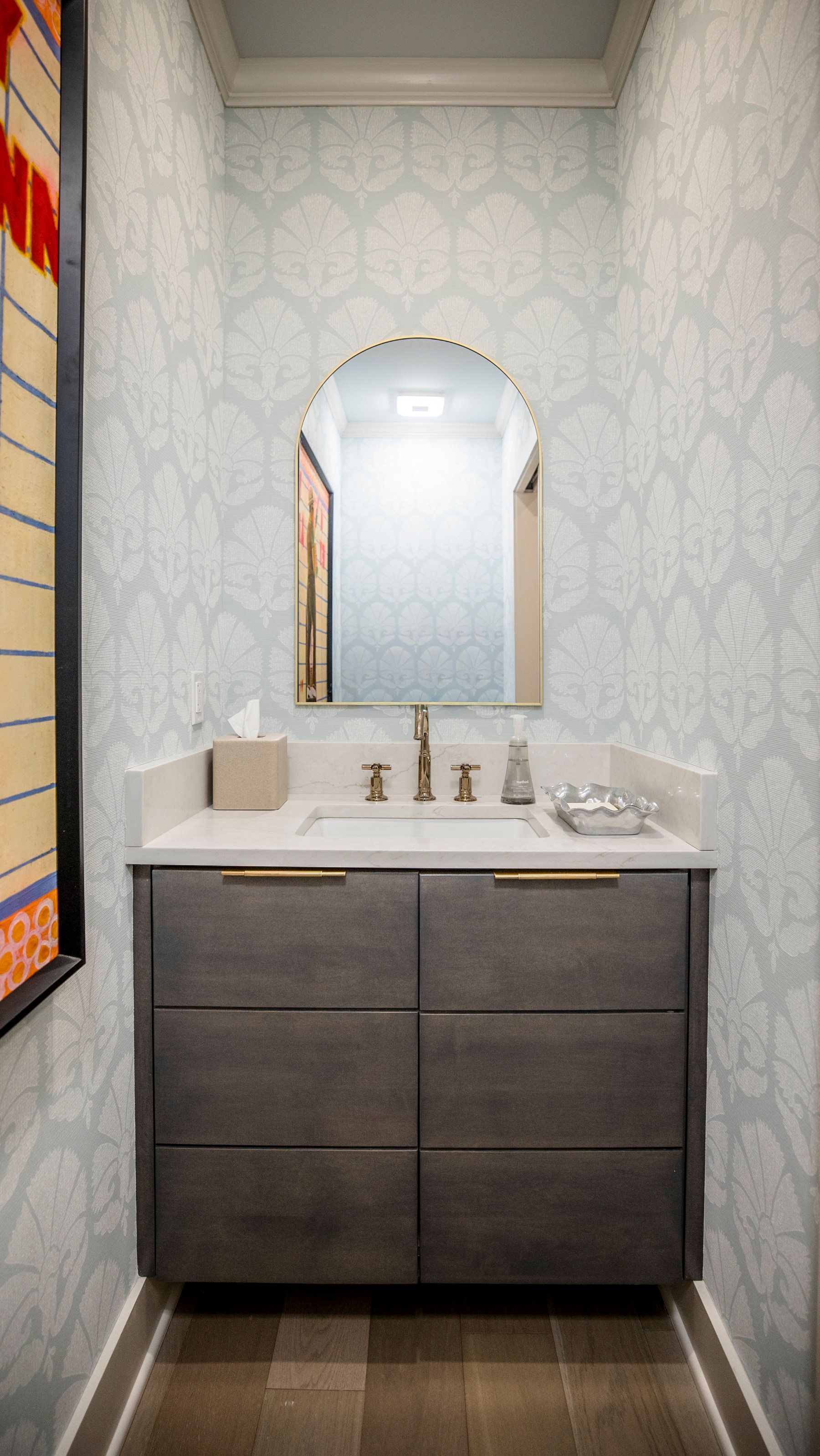 Small bathroom vanity with dark cabinets, white sink, arched mirror, and patterned wallpaper