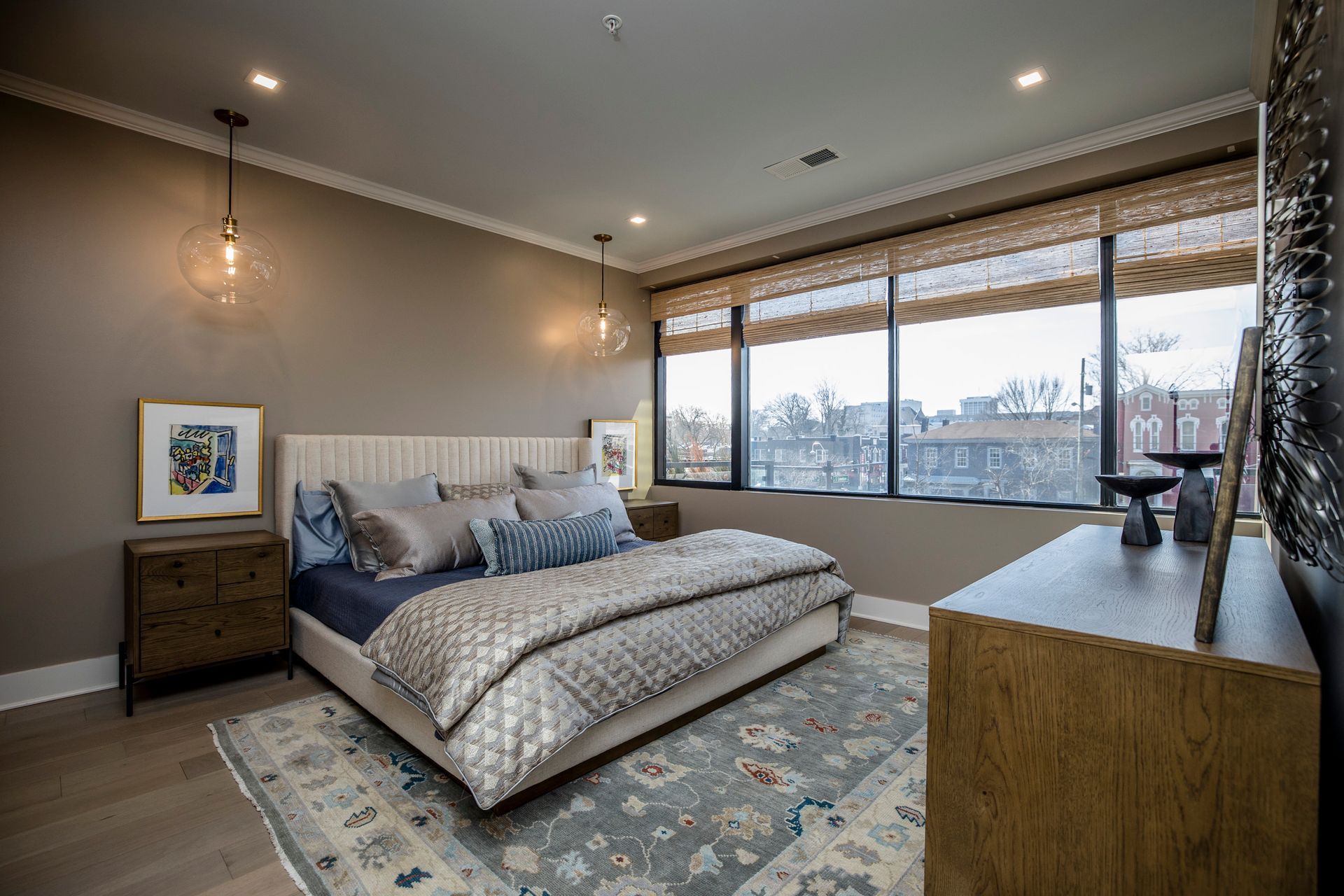 Modern bedroom with a large window, gray bed, patterned rug, and city view
