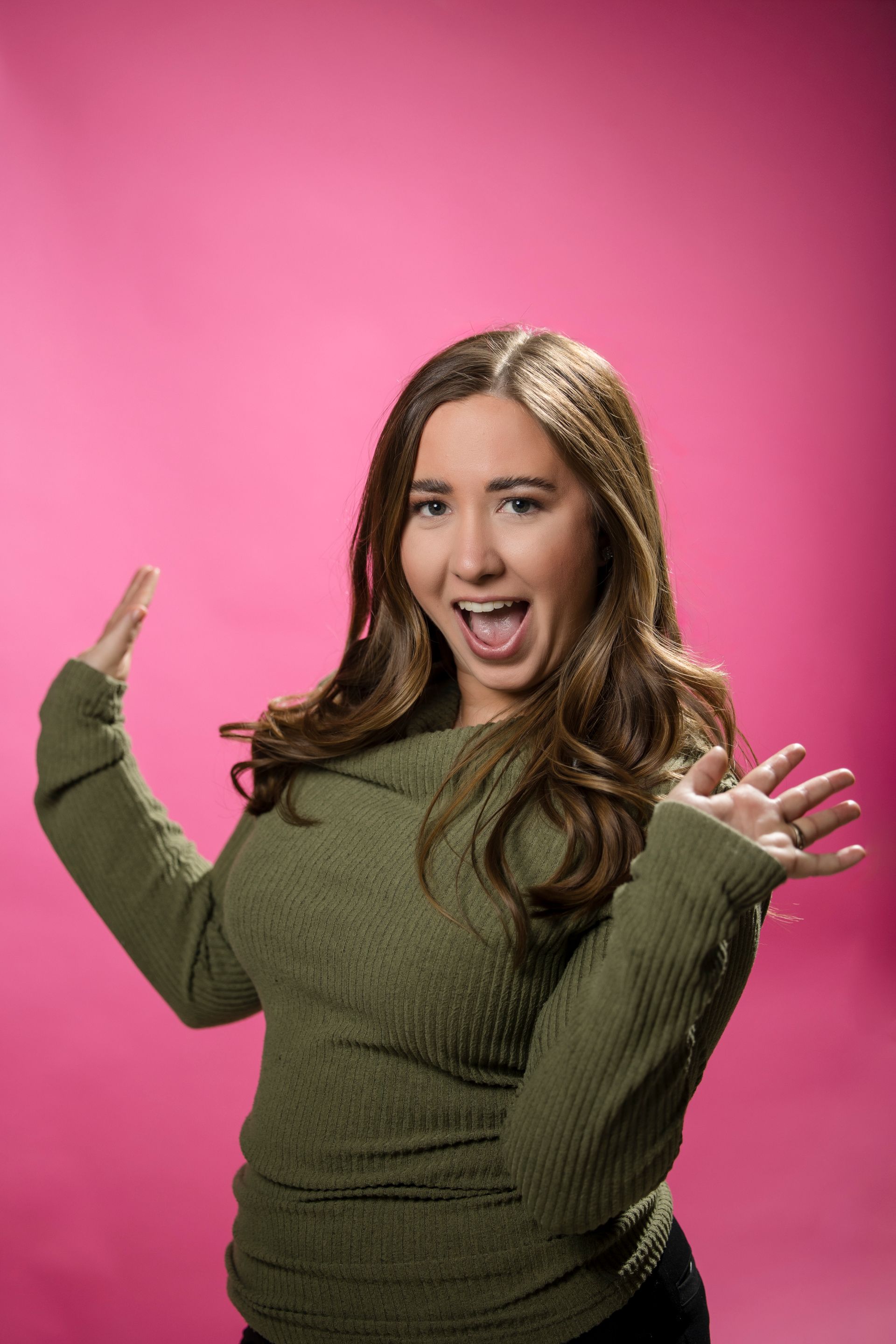 Smiling person in an olive sweater poses against a pink backdrop with hands raised.