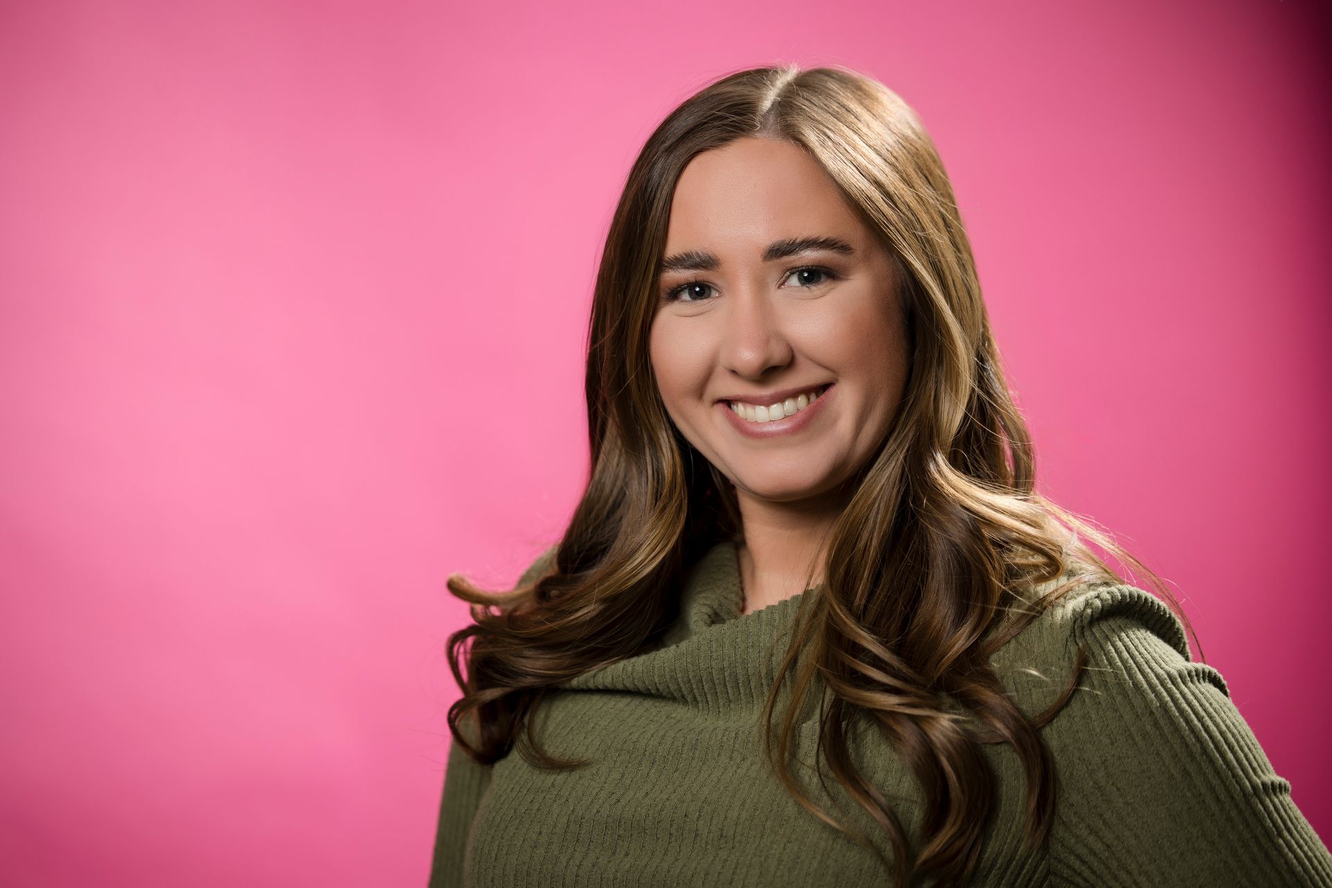 Smiling person in an olive green sweater against a bright pink background
