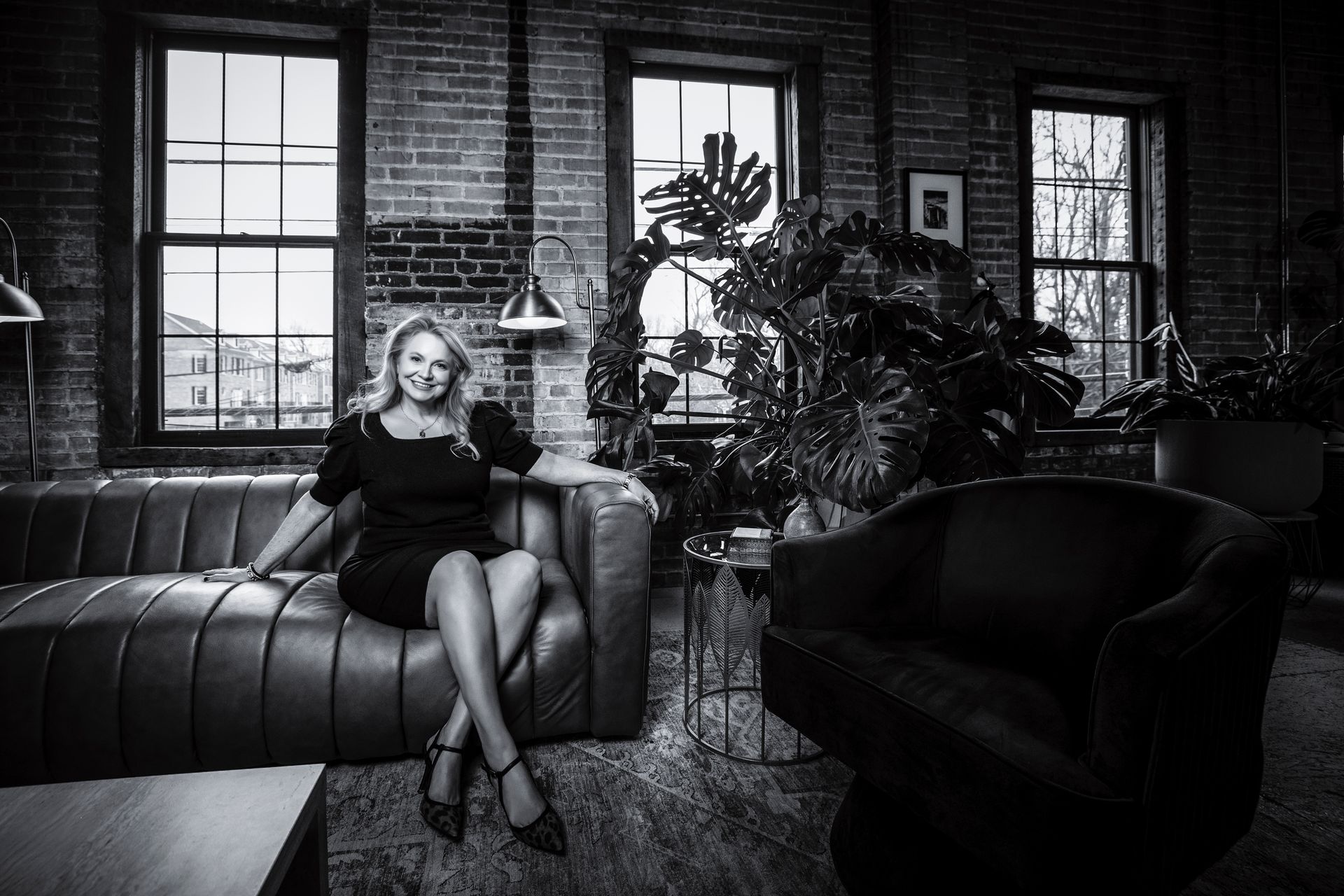 Woman seated on a leather sofa in a moody black-and-white loft with plants and large windows