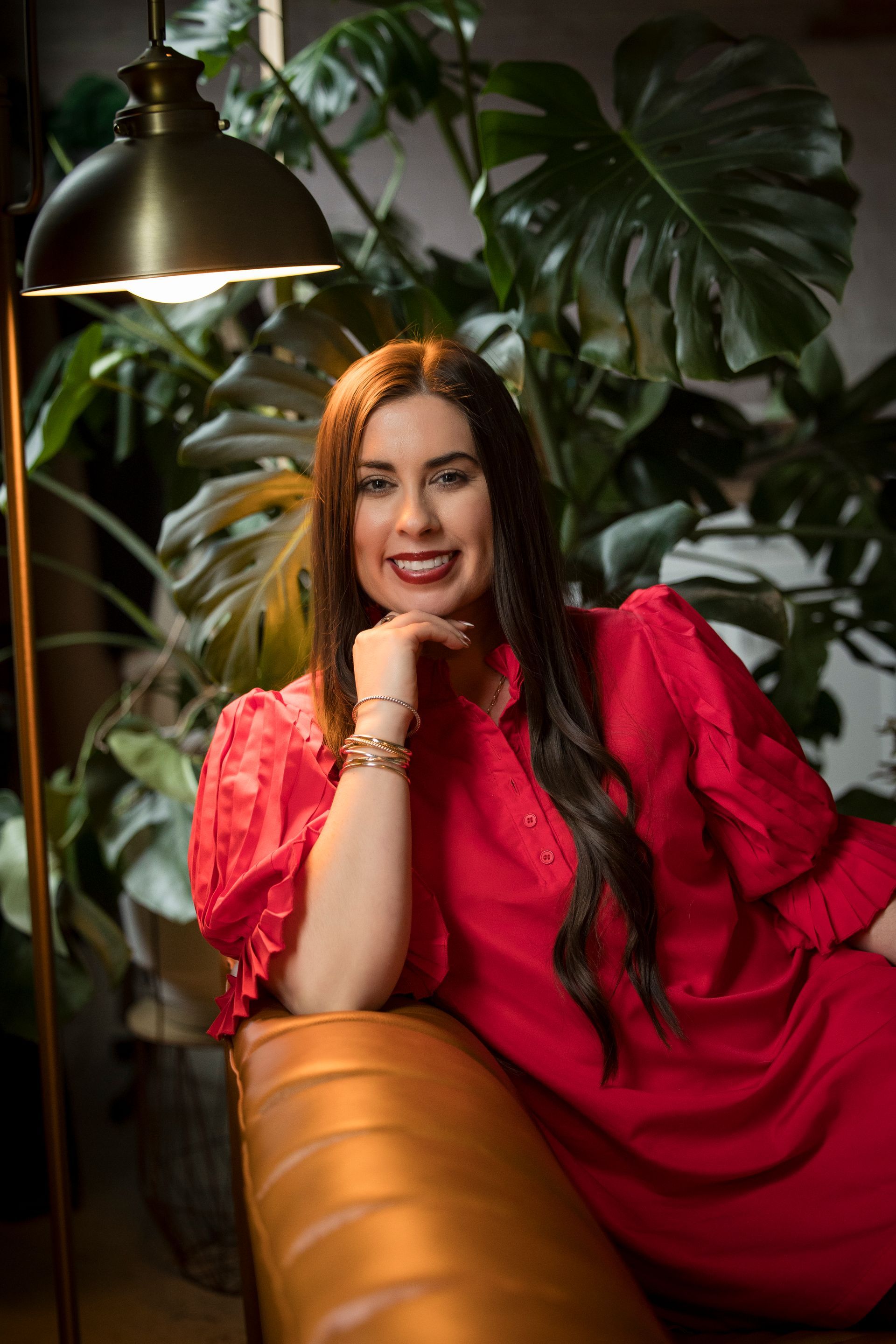 Woman in a bright red dress smiling on a leather sofa beside lush green plants and a hanging lamp