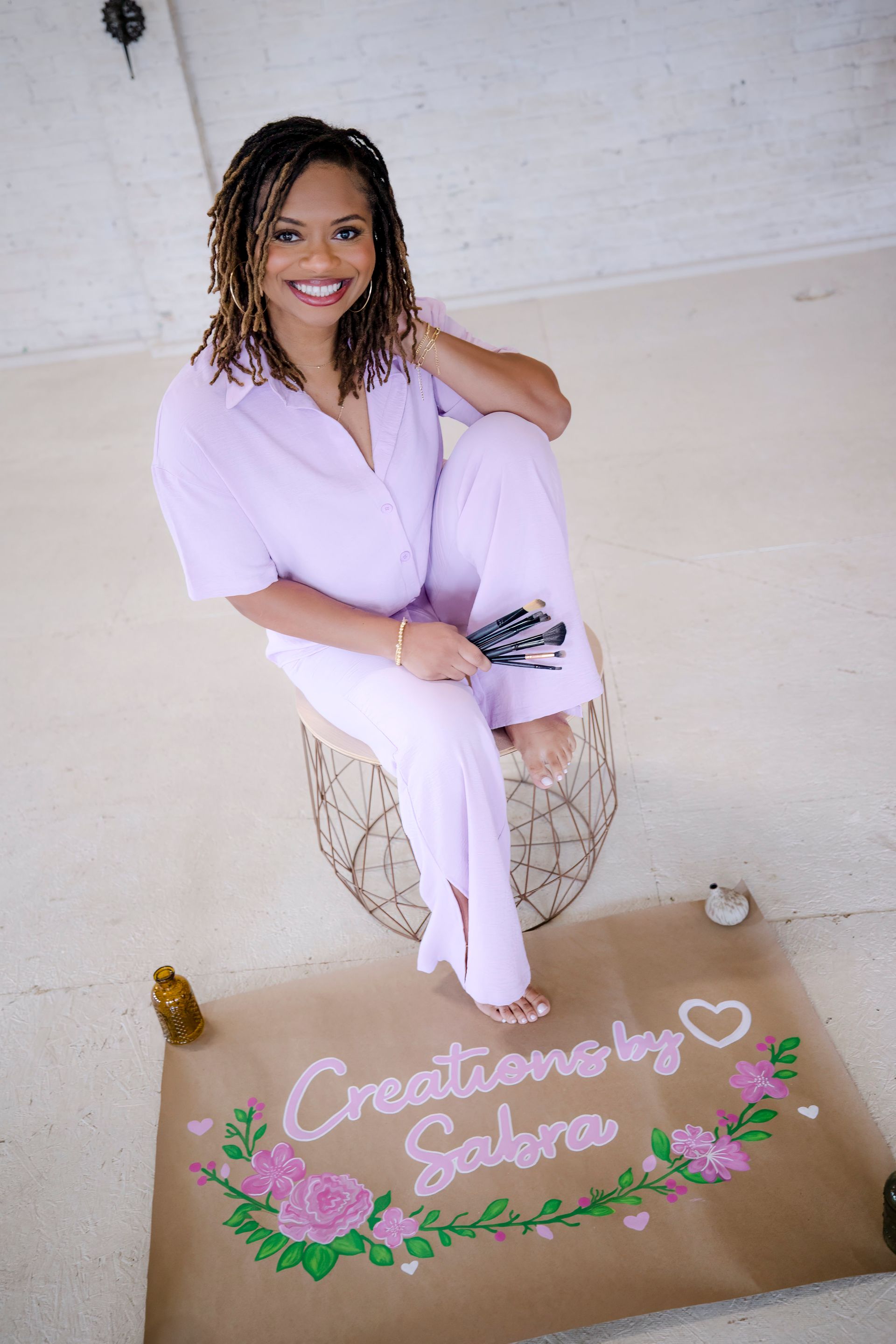Woman in lavender outfit sitting on a decorated sign with flowers and the words “Christiane’s 50,” smiling at the camera