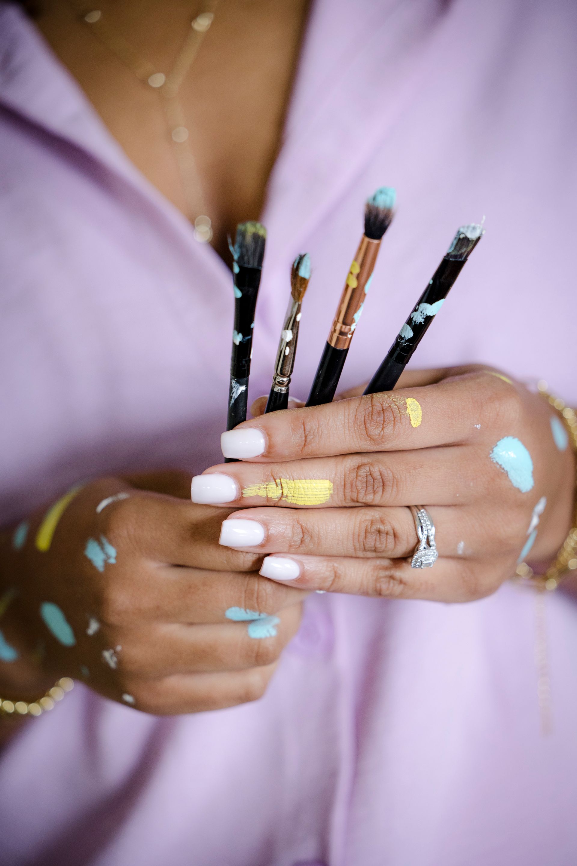 Hands holding paintbrushes, with colorful paint spots on fingers and a lavender shirt in the background