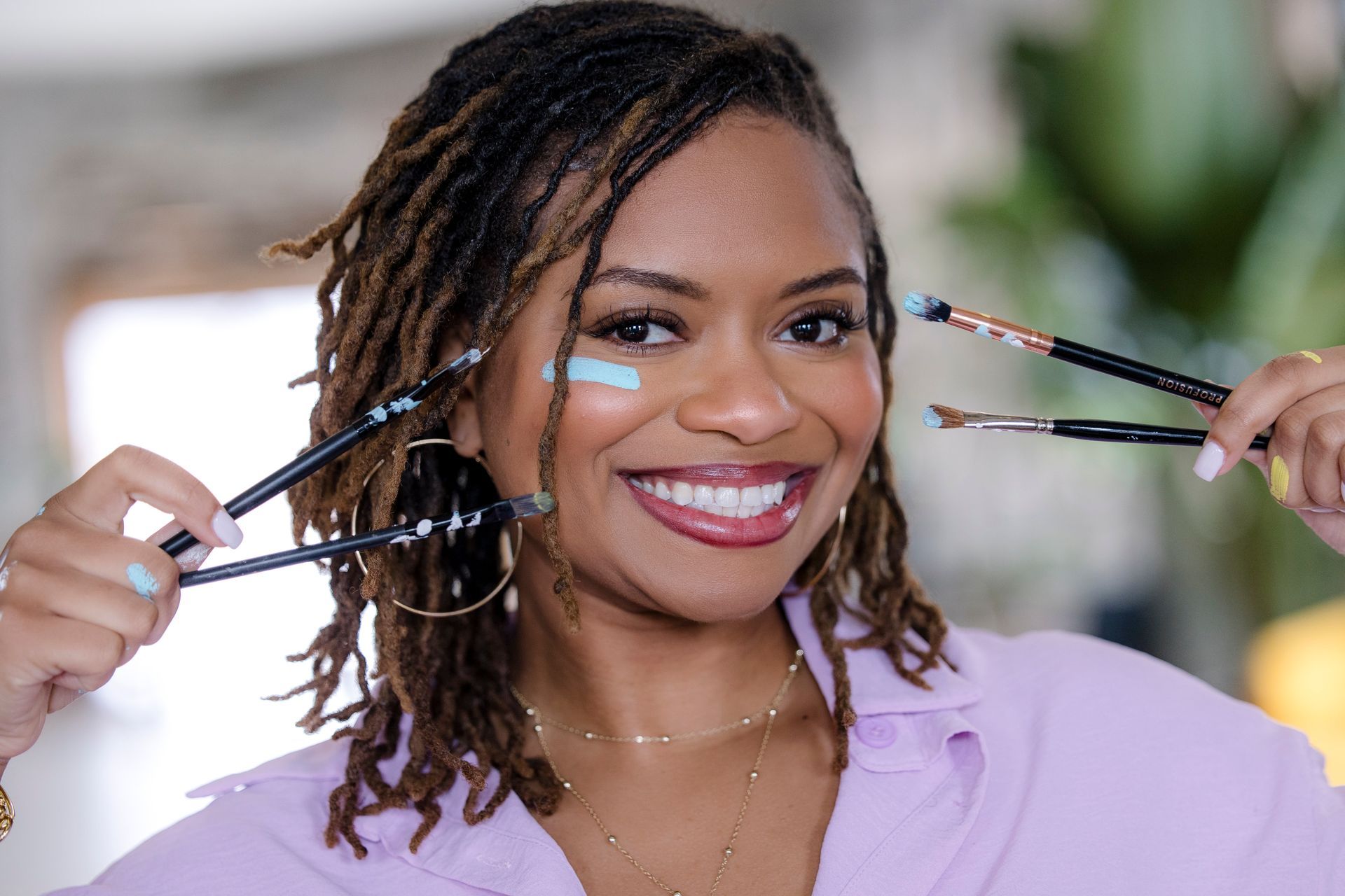Person smiling while holding multiple makeup brushes near their face in a bright room
