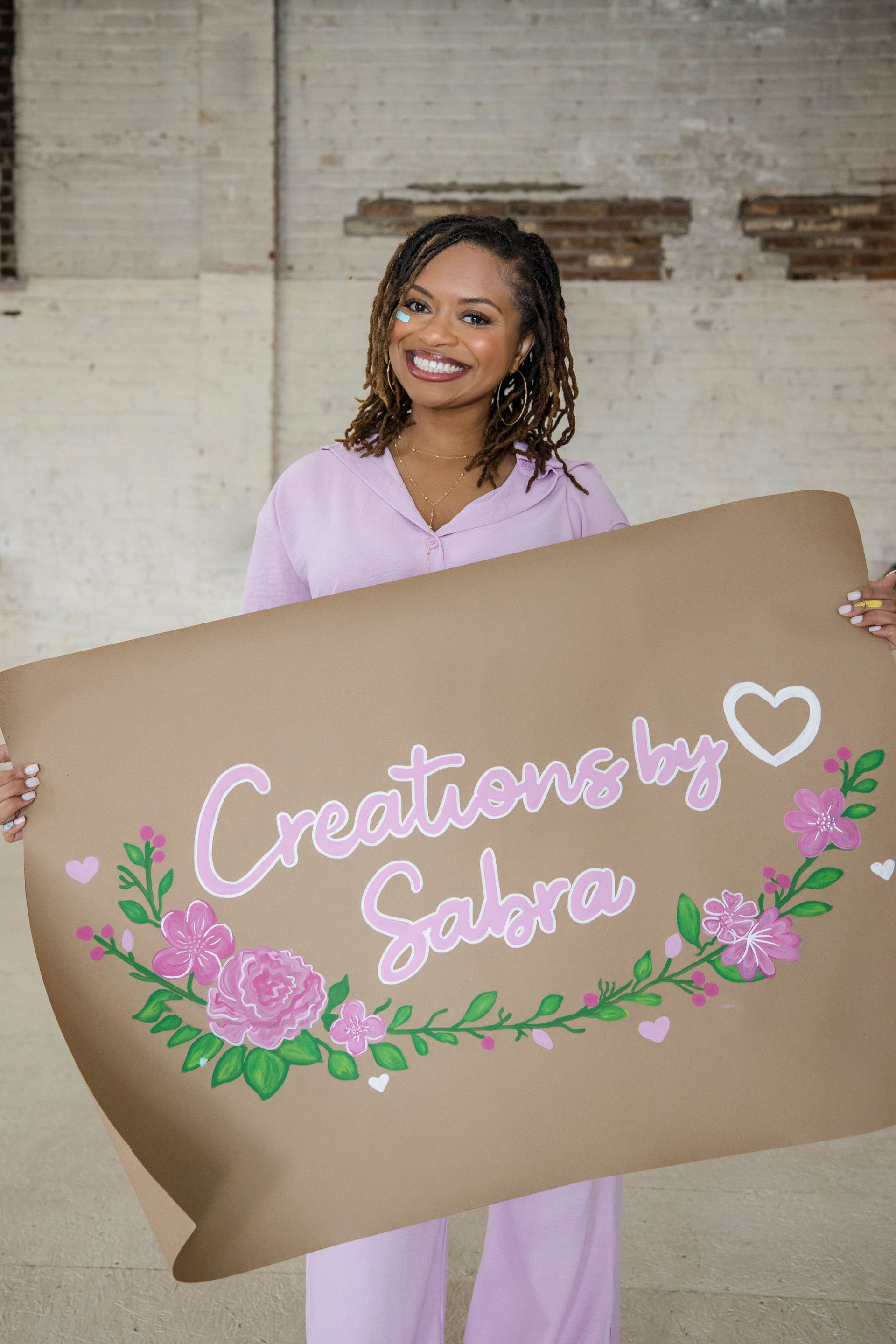 Woman holding a large brown sign reading “Creations by Sabrina” with pink floral decorations.