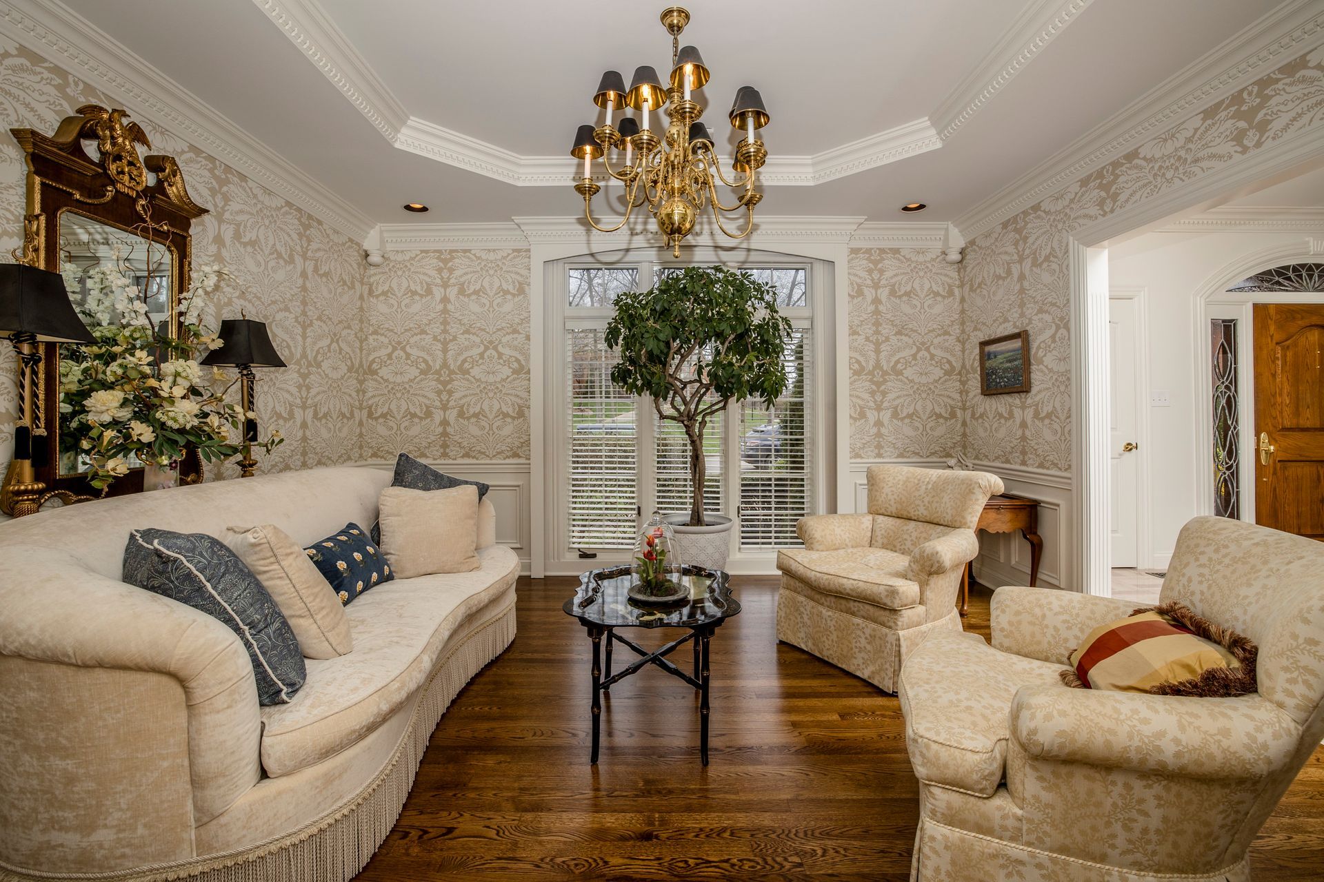 Elegant living room with cream sofas, chandelier, dark wood floor, and a large potted tree by tall windows
