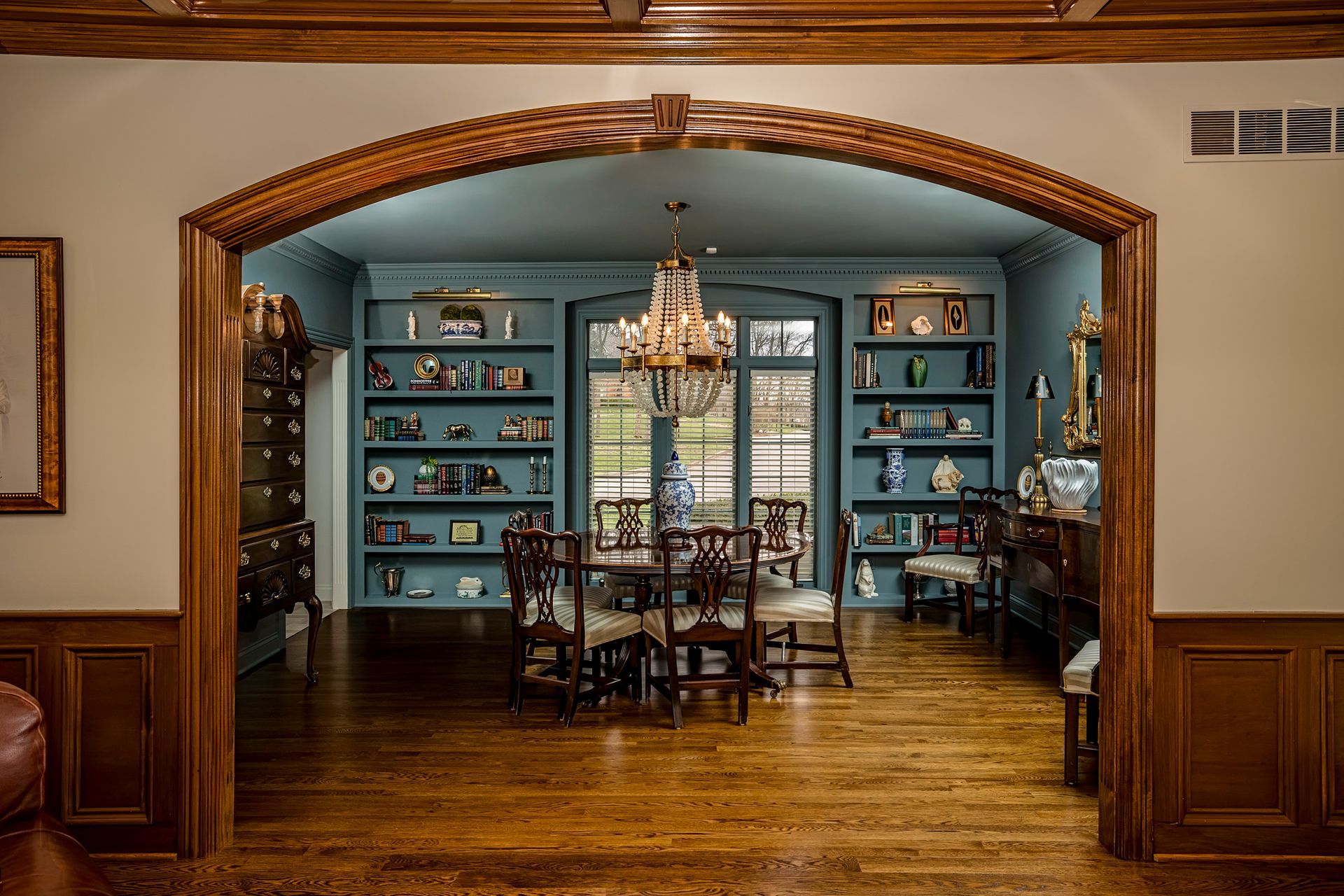 Dining room seen through an arched opening, with blue built-in shelves, chandelier, and hardwood floors.