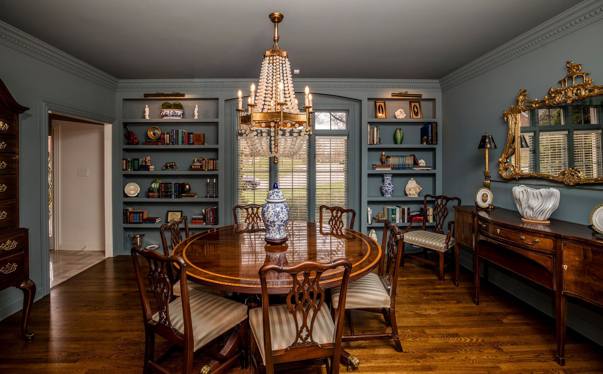 Elegant dining room with chandelier, wooden table, blue built-in shelves, and ornate mirror.