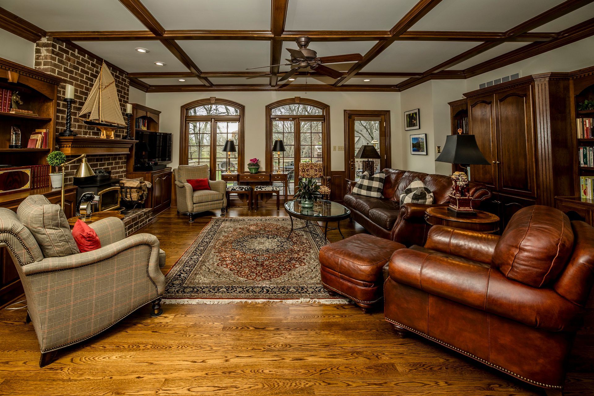 Warm living room with leather sofa, armchairs, patterned rug, fireplace, wood floors, and large windows
