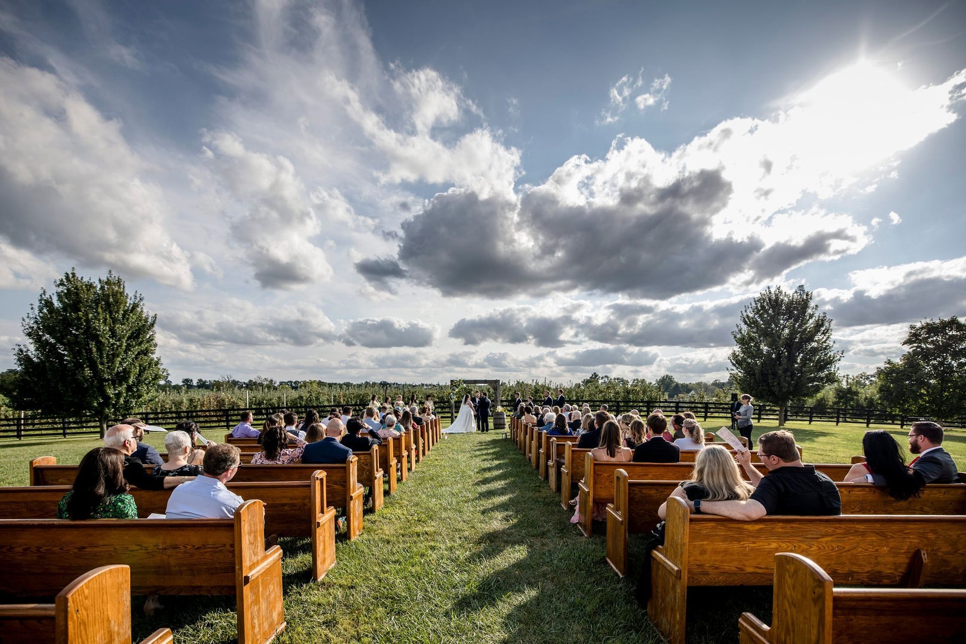 Wedding ceremony outdoors with rows of guests seated, sunny sky, and couple at altar.