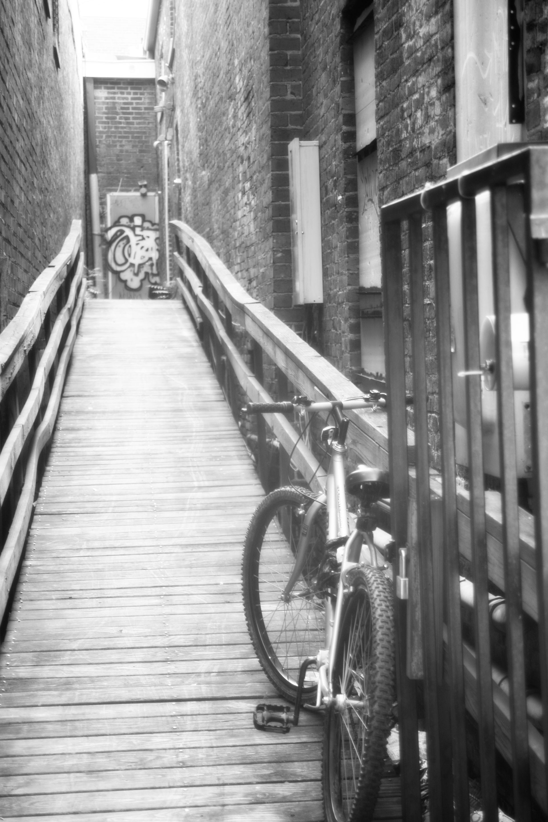 Black-and-white alley with a bicycle leaning beside a railing on a narrow wooden ramp.