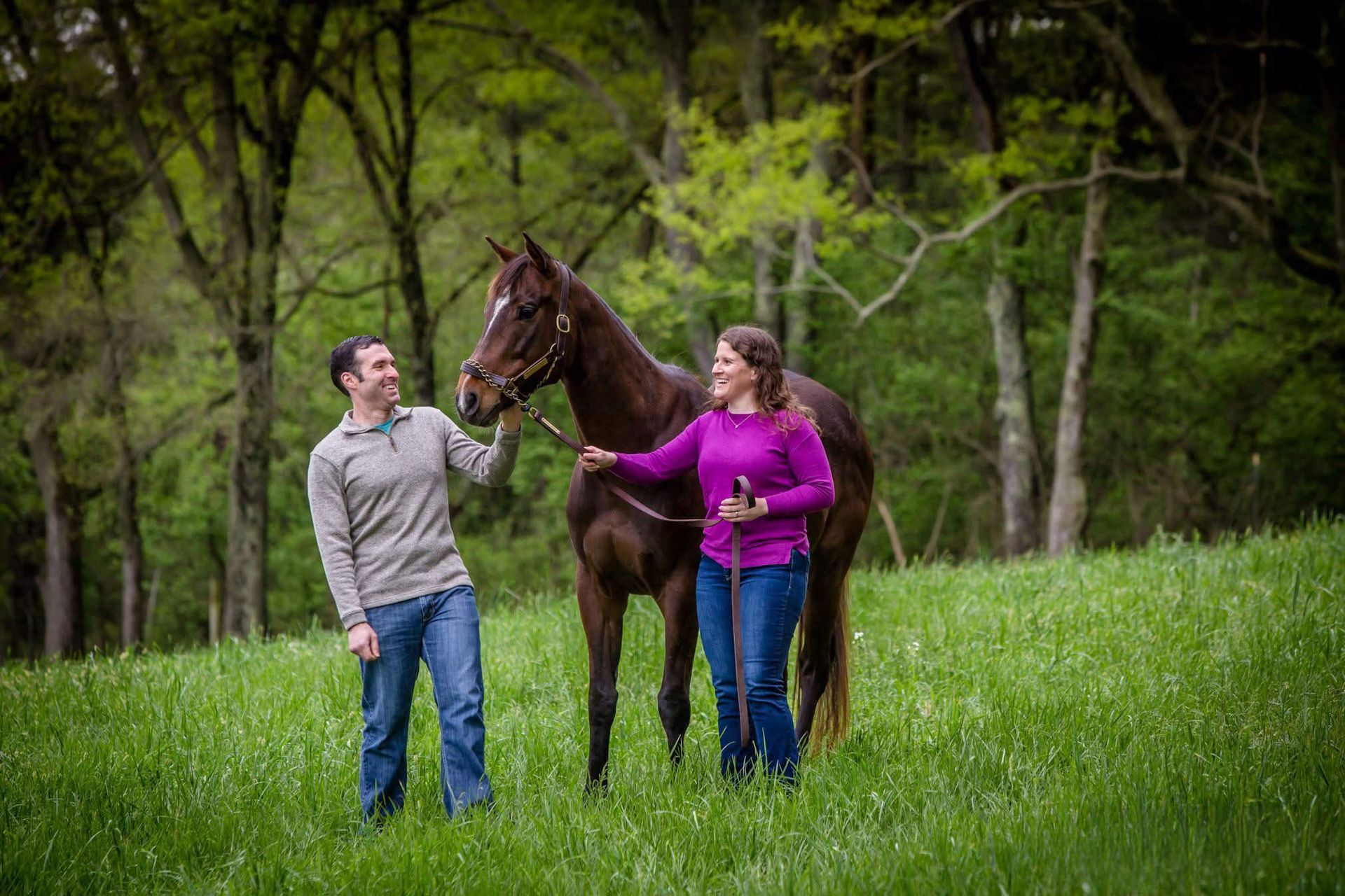 Two people standing with a brown horse in a green field, one holding the reins.