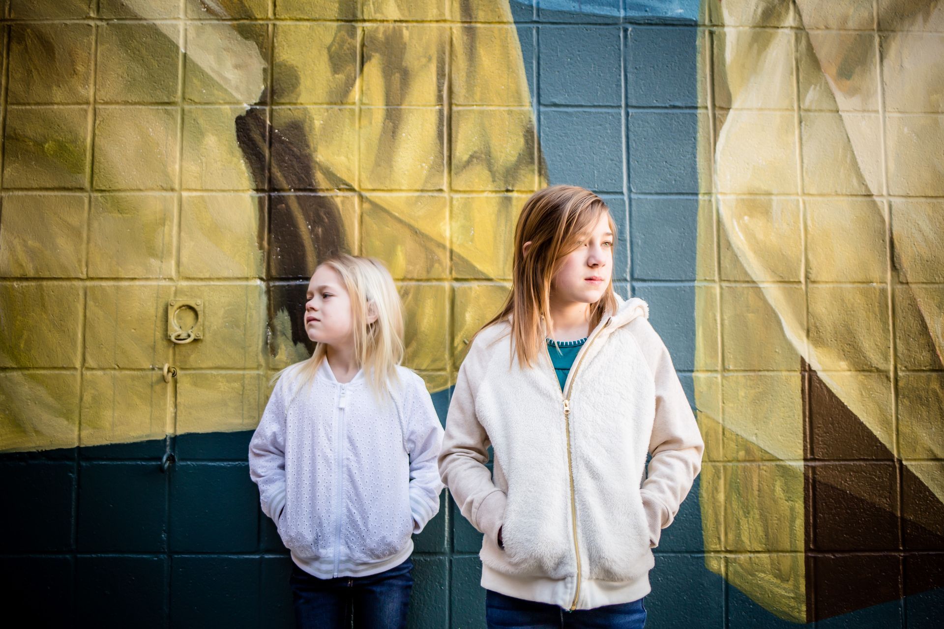 Two children in white jackets stand against a colorful tiled mural, looking off to the side.