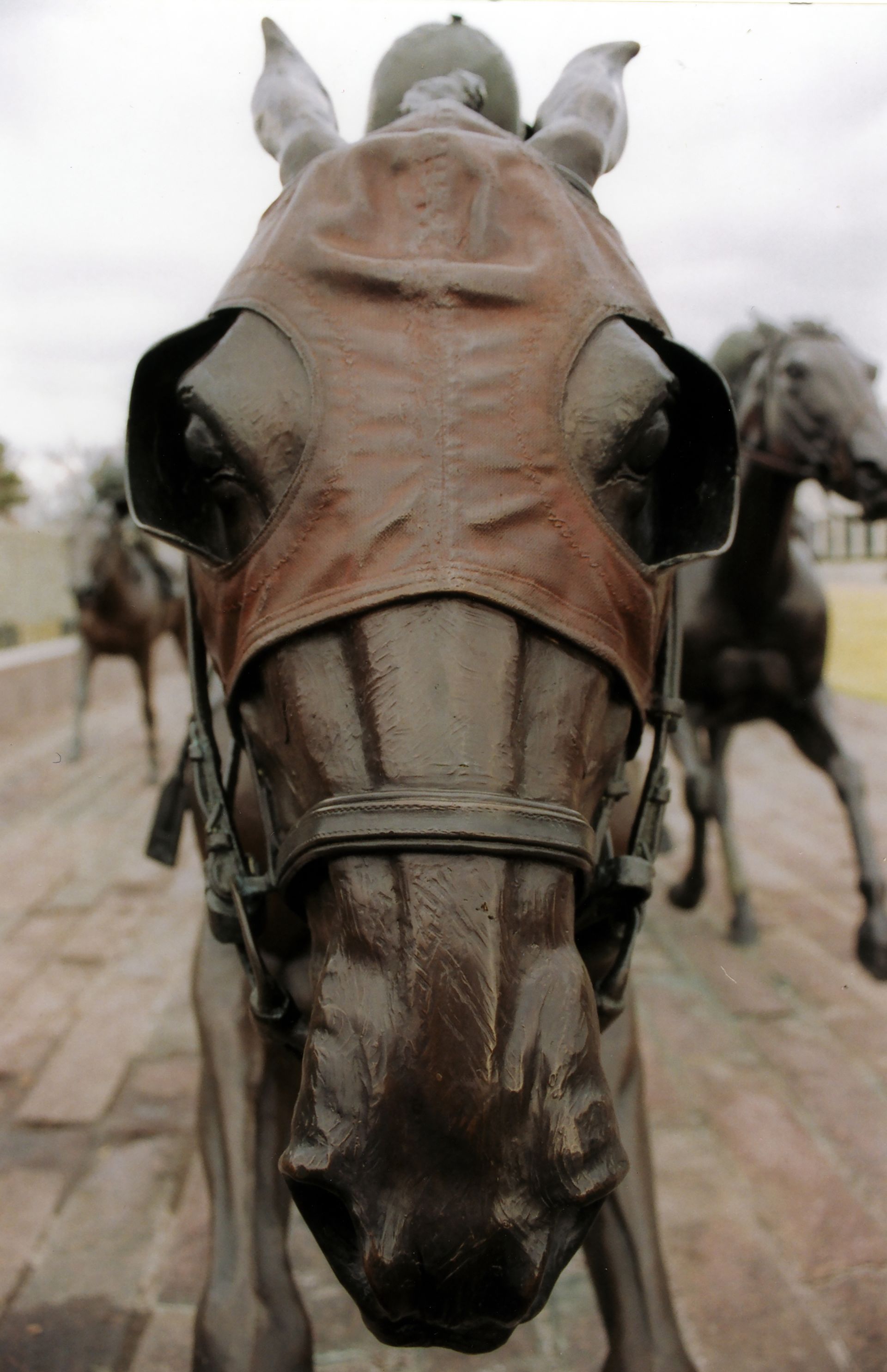 Close-up of a horse statue wearing a leather-like mask, with another horse statue blurred in the background.