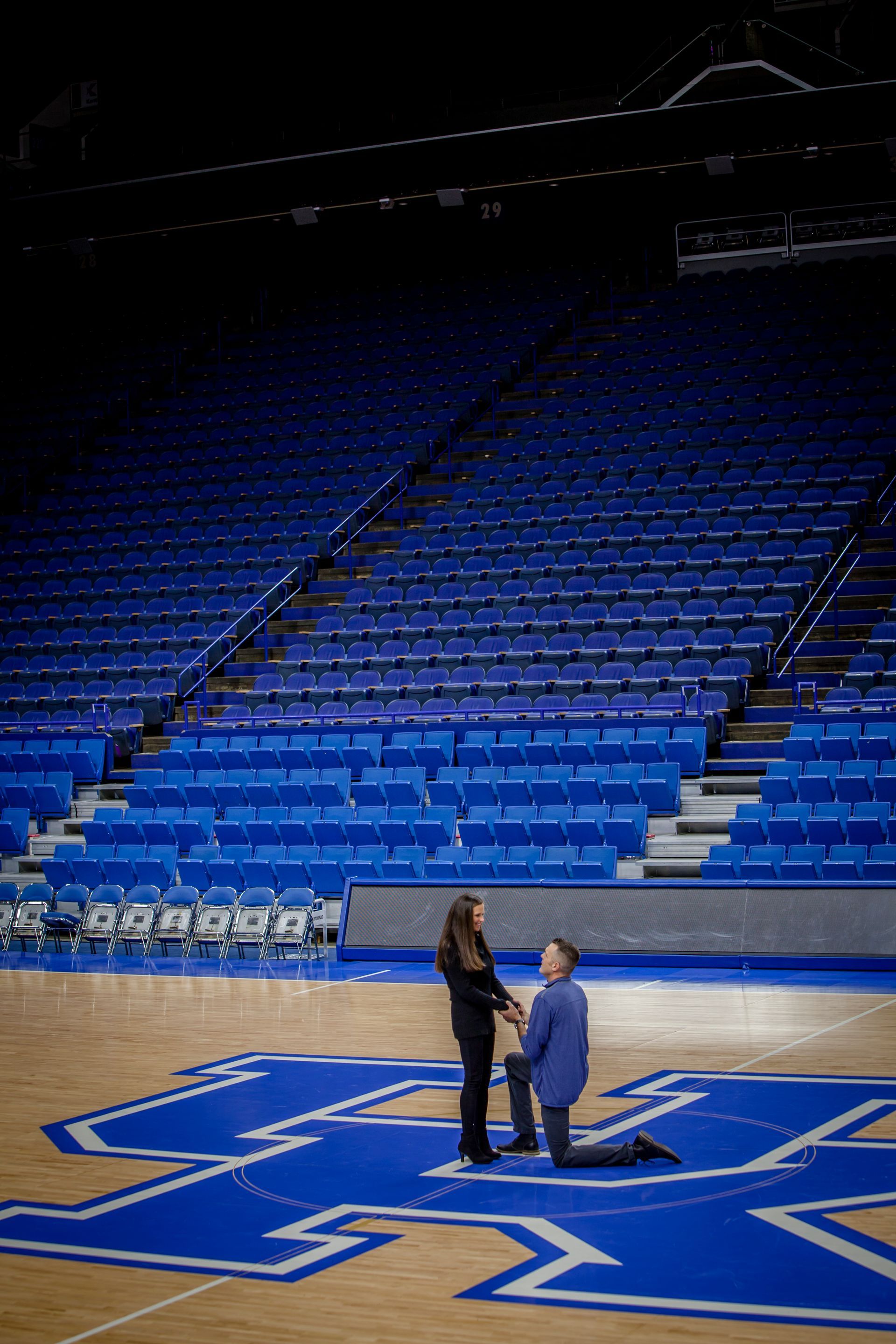 Two people standing on a blue basketball court inside an empty arena with blue seats