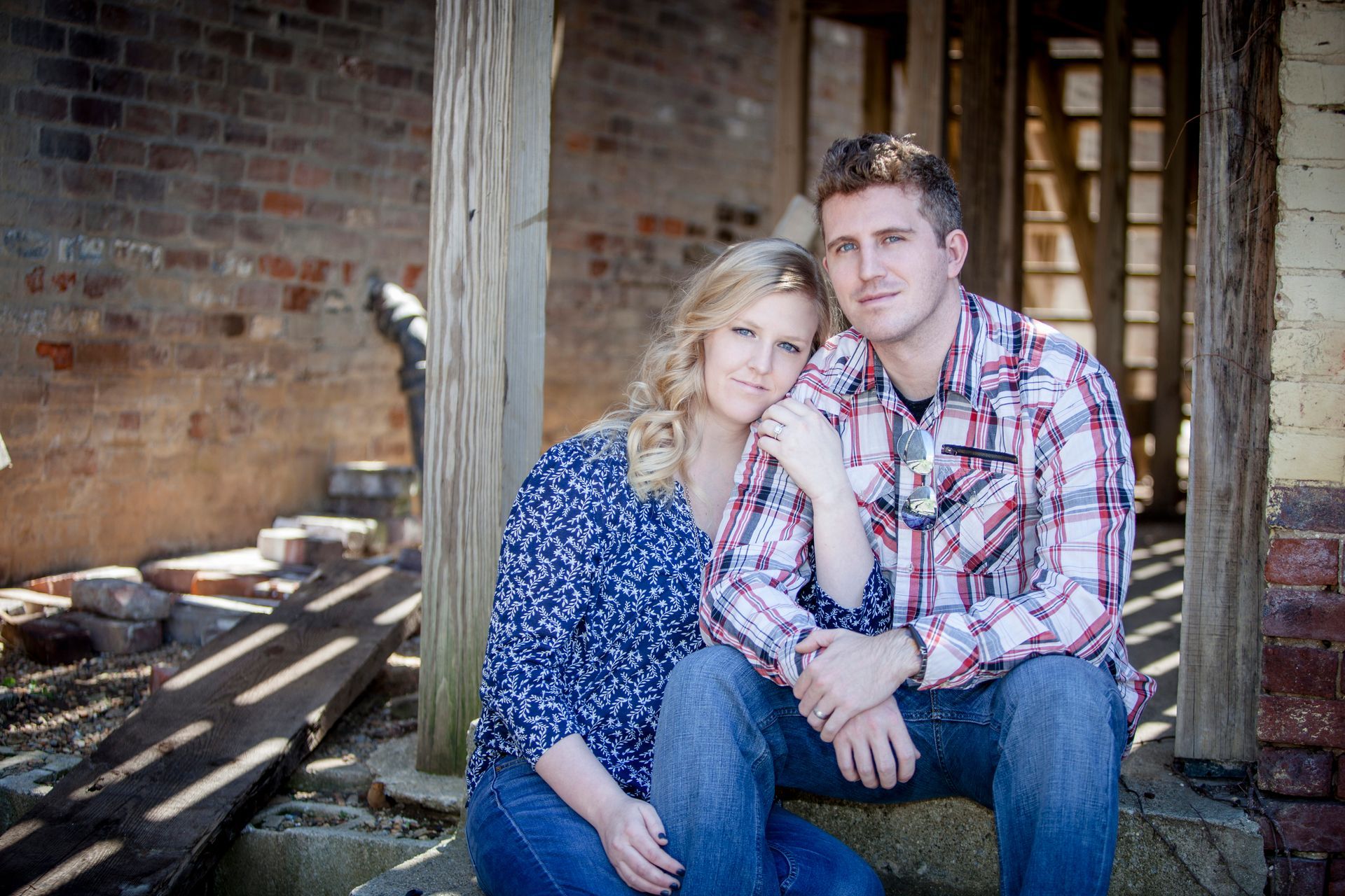 Couple sitting together on a wooden porch, wearing blue and plaid, with a rustic brick wall behind them