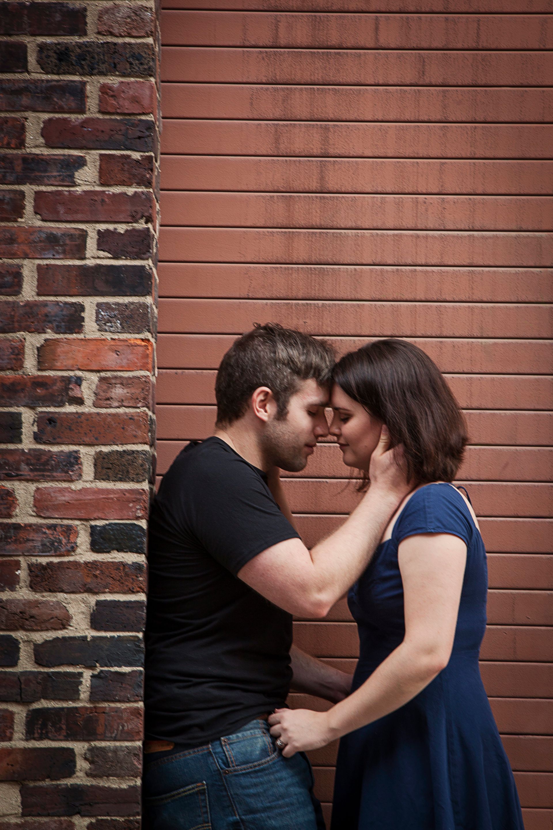 Couple embracing against a brick wall, the woman in a blue dress and the man in a black shirt.