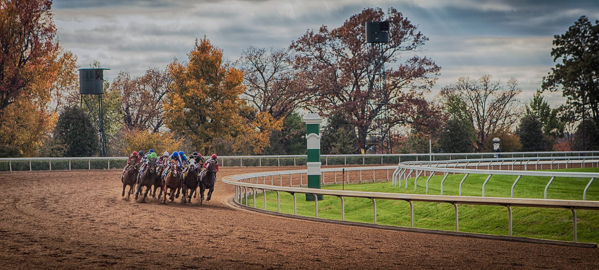 Jockeys on horses rounding a dirt track bend with white railings and autumn trees in the background
