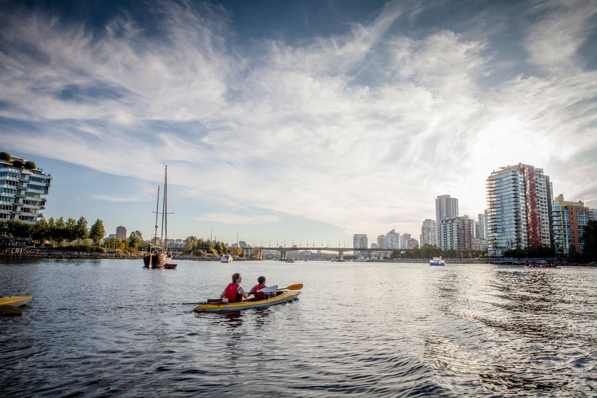 Kayak with two people on a wide river, surrounded by city buildings under a cloudy sky