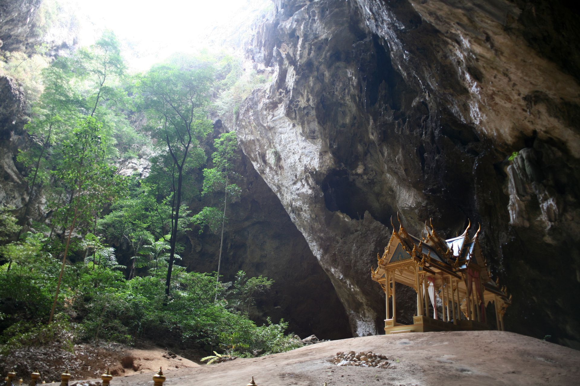 Small golden shrine under a large cave overhang, surrounded by lush green forest and rocky ground.
