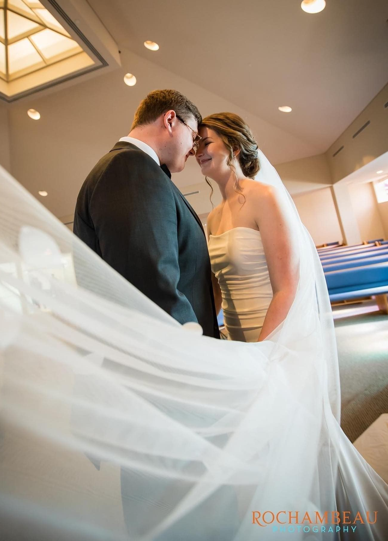 Bride and groom embracing, veil billowing, in a church. Bride wears strapless dress, groom in suit.