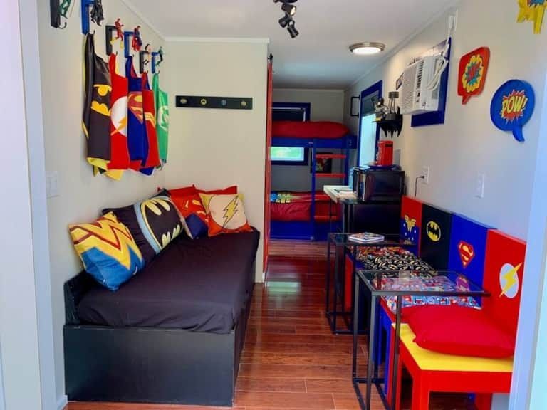 Bright, narrow bedroom with colorful superhero decor, black daybed, red desk, and bunk bed in the back.