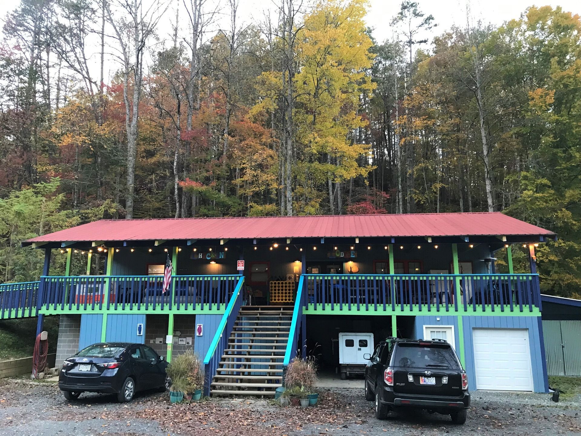 Colorful stilt house with red roof and cars parked below, set against autumn trees
