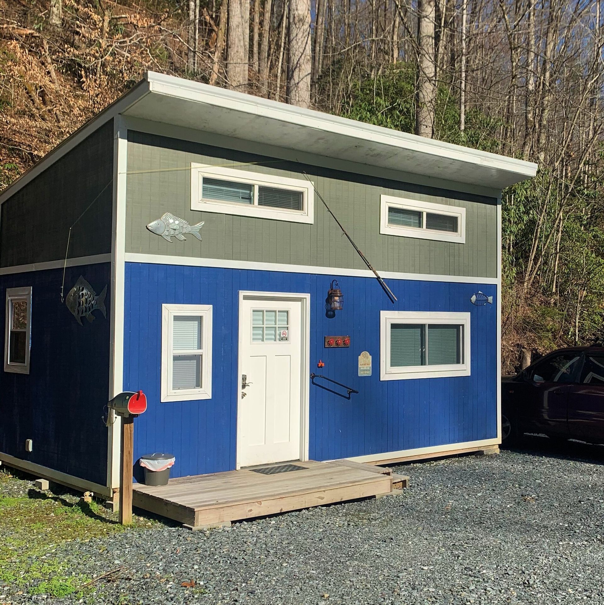 Small blue-and-gray cabin with white door and windows on a gravel lot in the woods