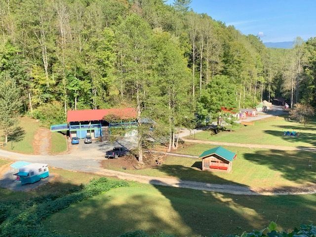 Rural campground with cabins, red-roof shelter, grassy field, and wooded hills under bright sunlight
