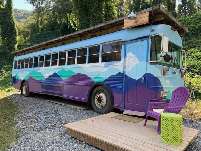 Colorful purple and blue bus parked beside a wooden deck with chairs in a wooded setting