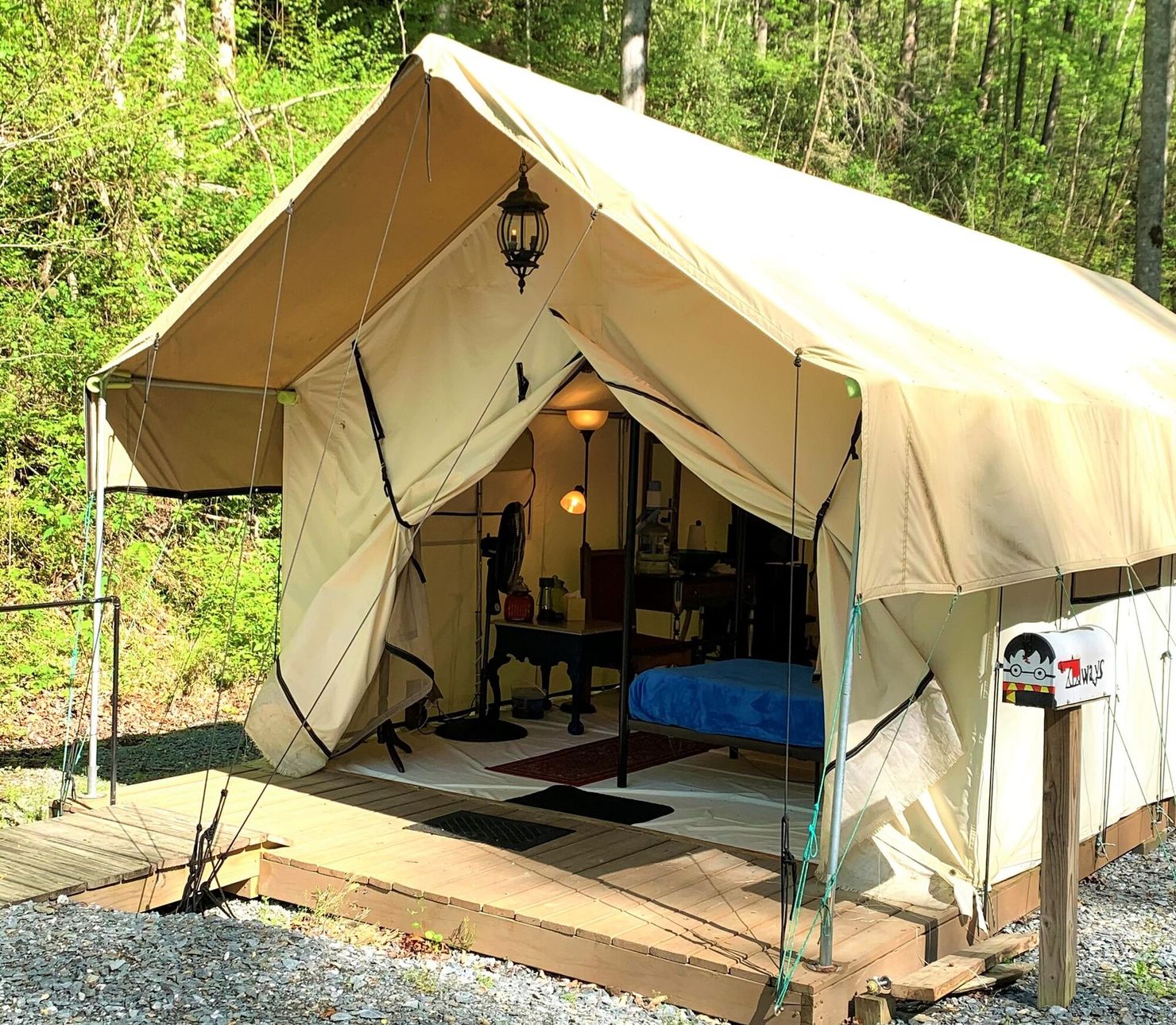 Beige canvas safari tent with open front on a forest campsite, with chairs and a lamp inside.