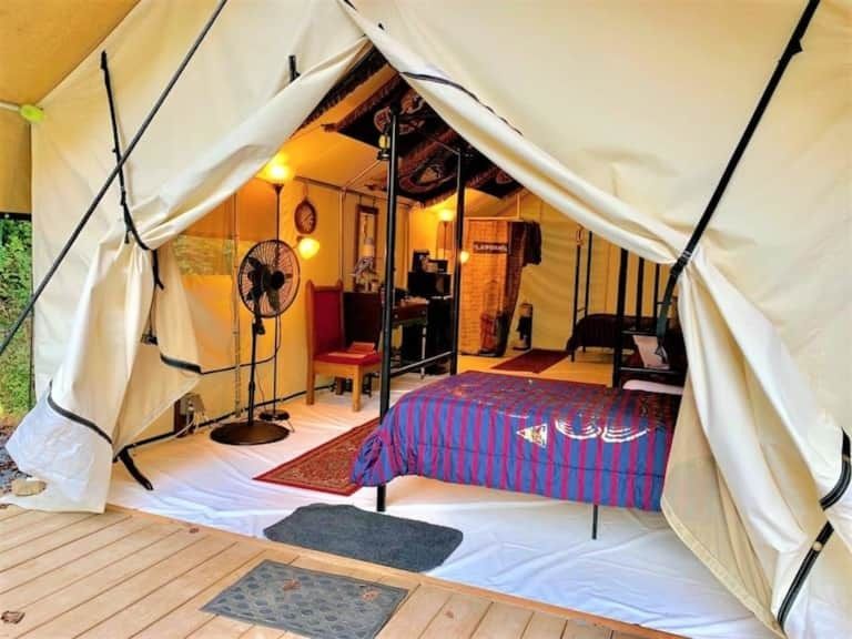 Interior of a white glamping tent with a bed, fan, rugs, and warm lighting
