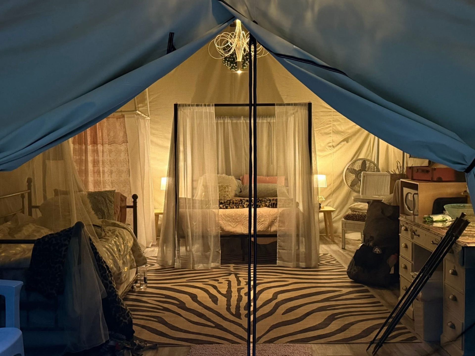 Inside a large tent, a cozy bed with sheer curtains sits on a zebra rug, lit warmly by lamps.