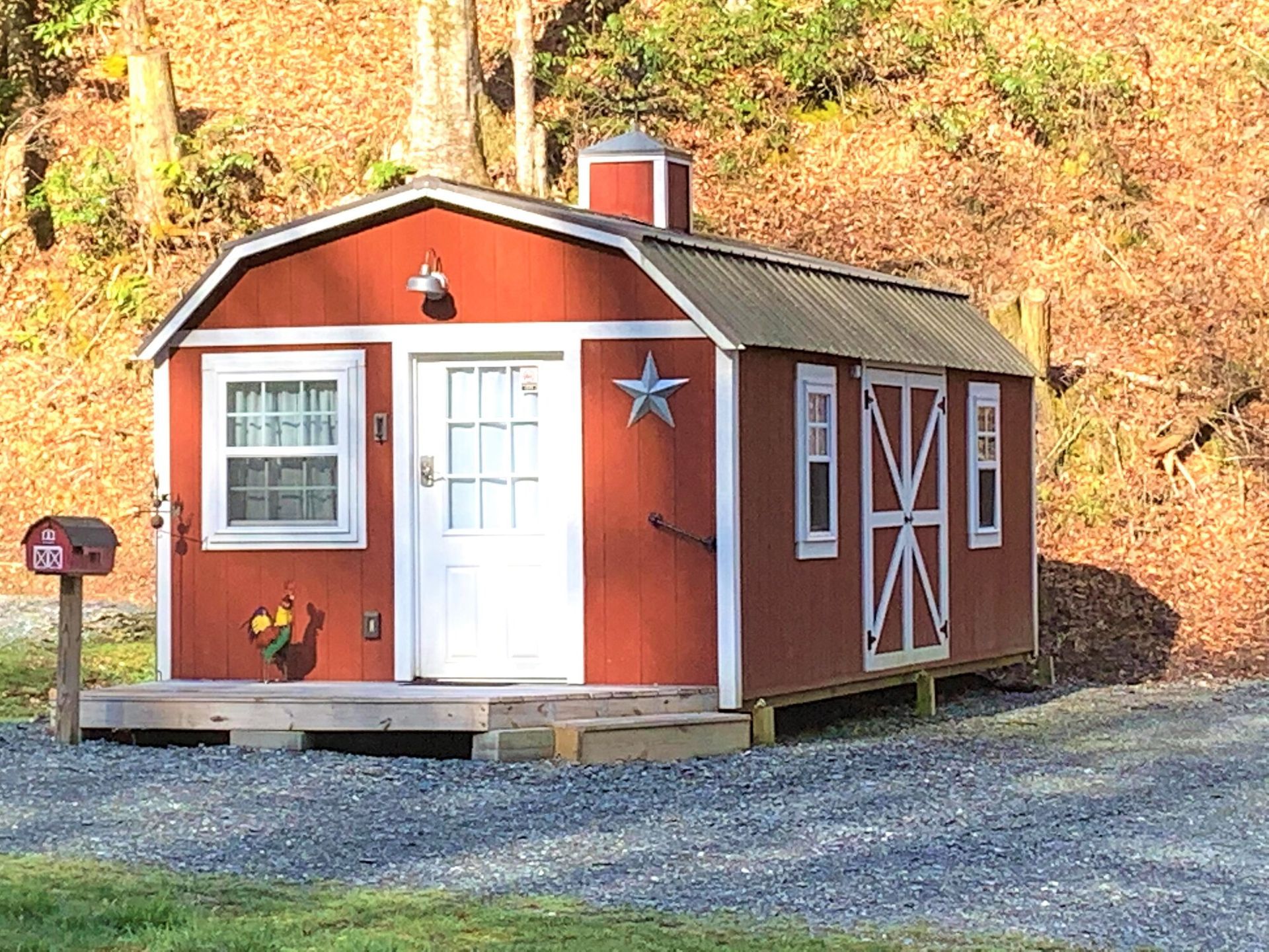Small red cabin with white trim and a star on the door, set on a gravel lot against a wooded hillside.