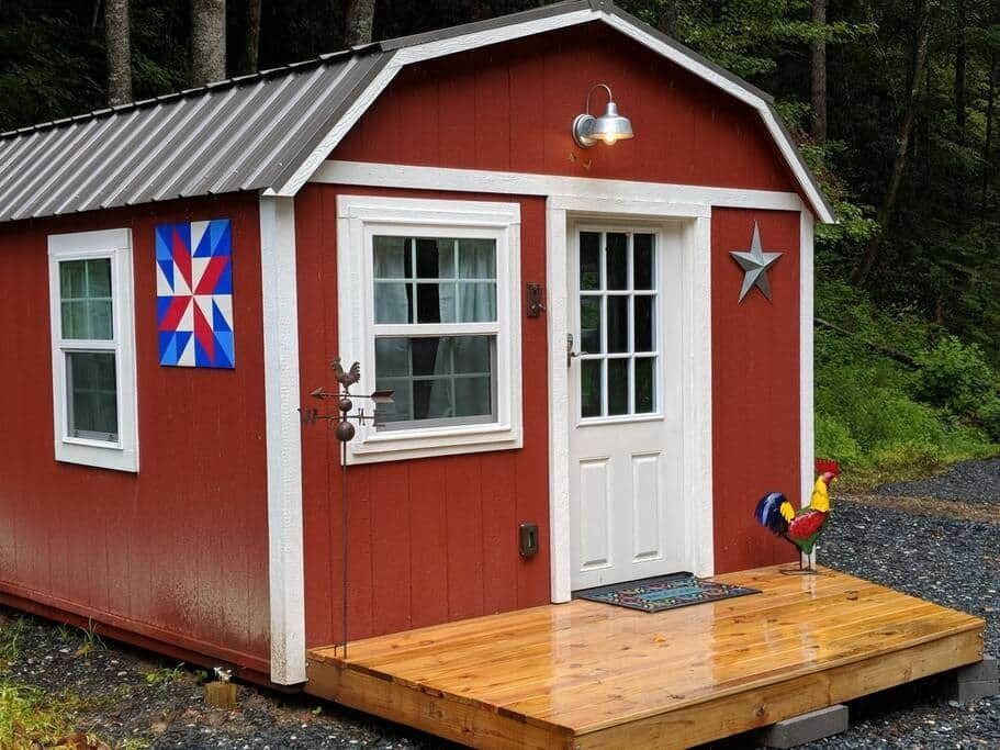 Small red cabin with white trim, porch, star decoration, and colorful quilt in a wooded area.
