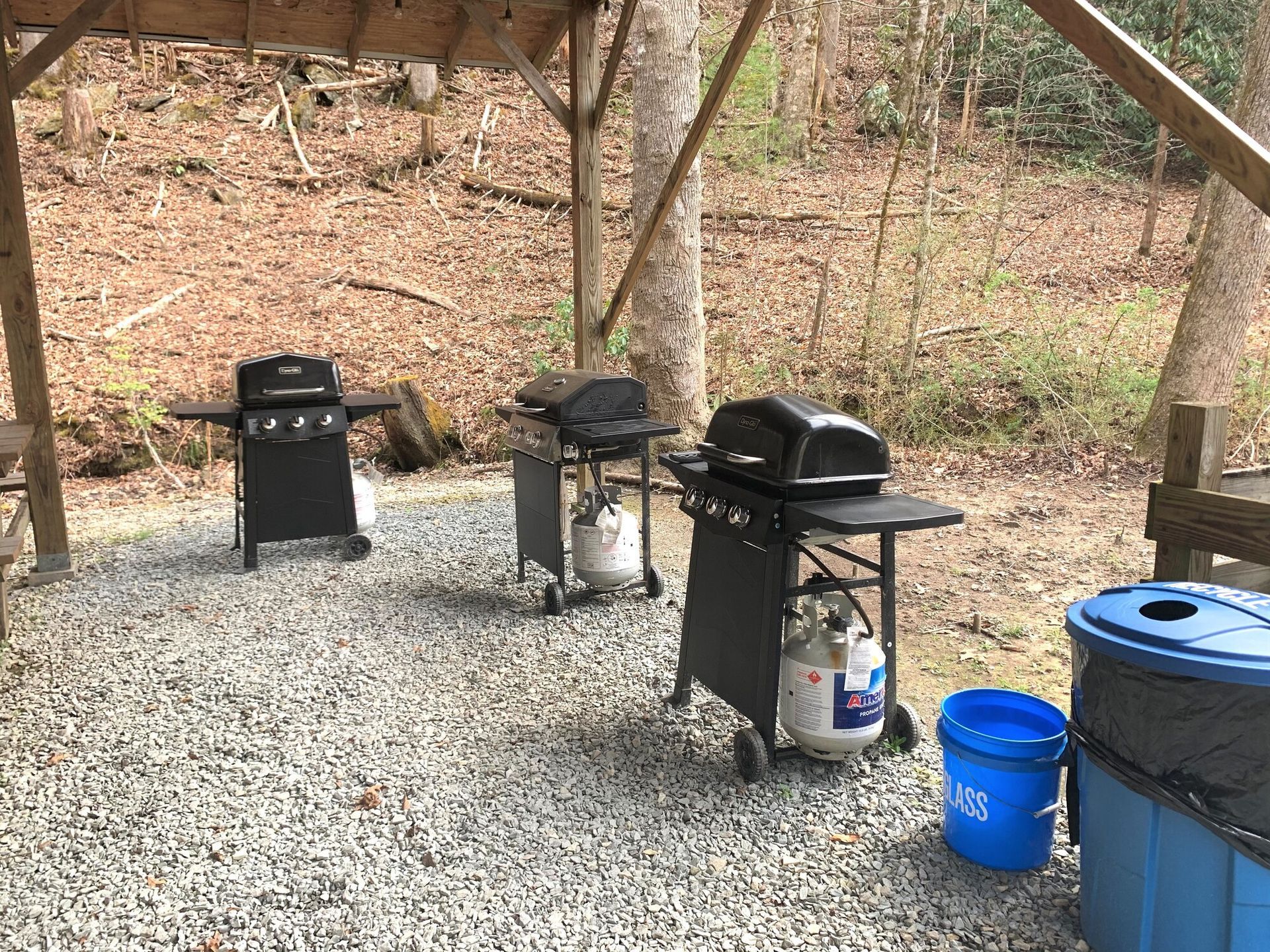Three black grills under a wooden shelter, with buckets and gravel ground.
