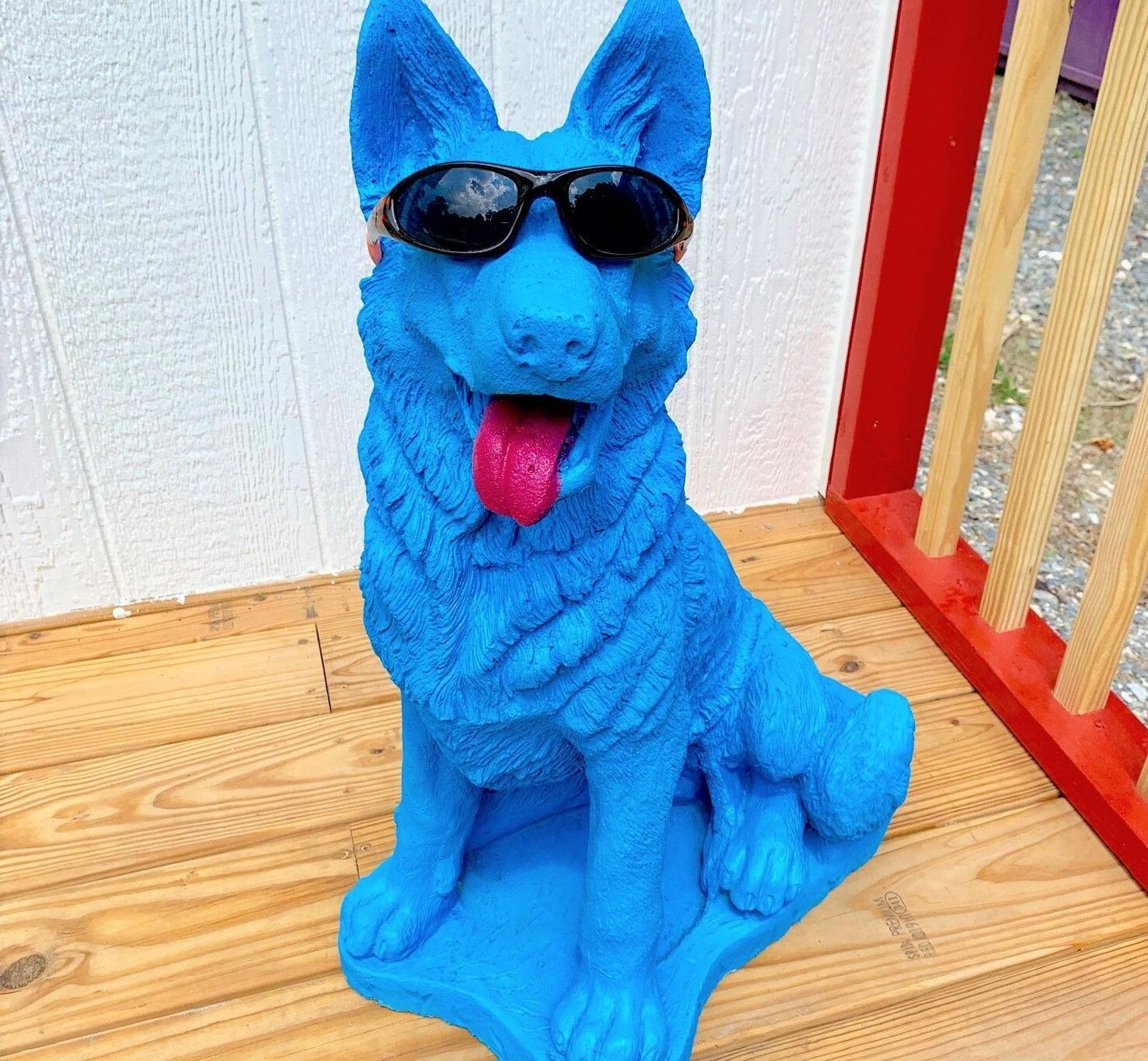 Blue dog statue wearing sunglasses with tongue out, sitting on a wooden deck near a red railing