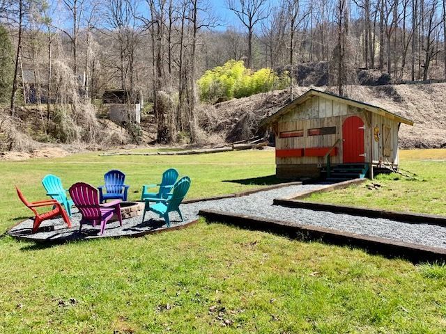 Colorful Adirondack chairs around a fire pit beside a small shed on a grassy lawn in a wooded park