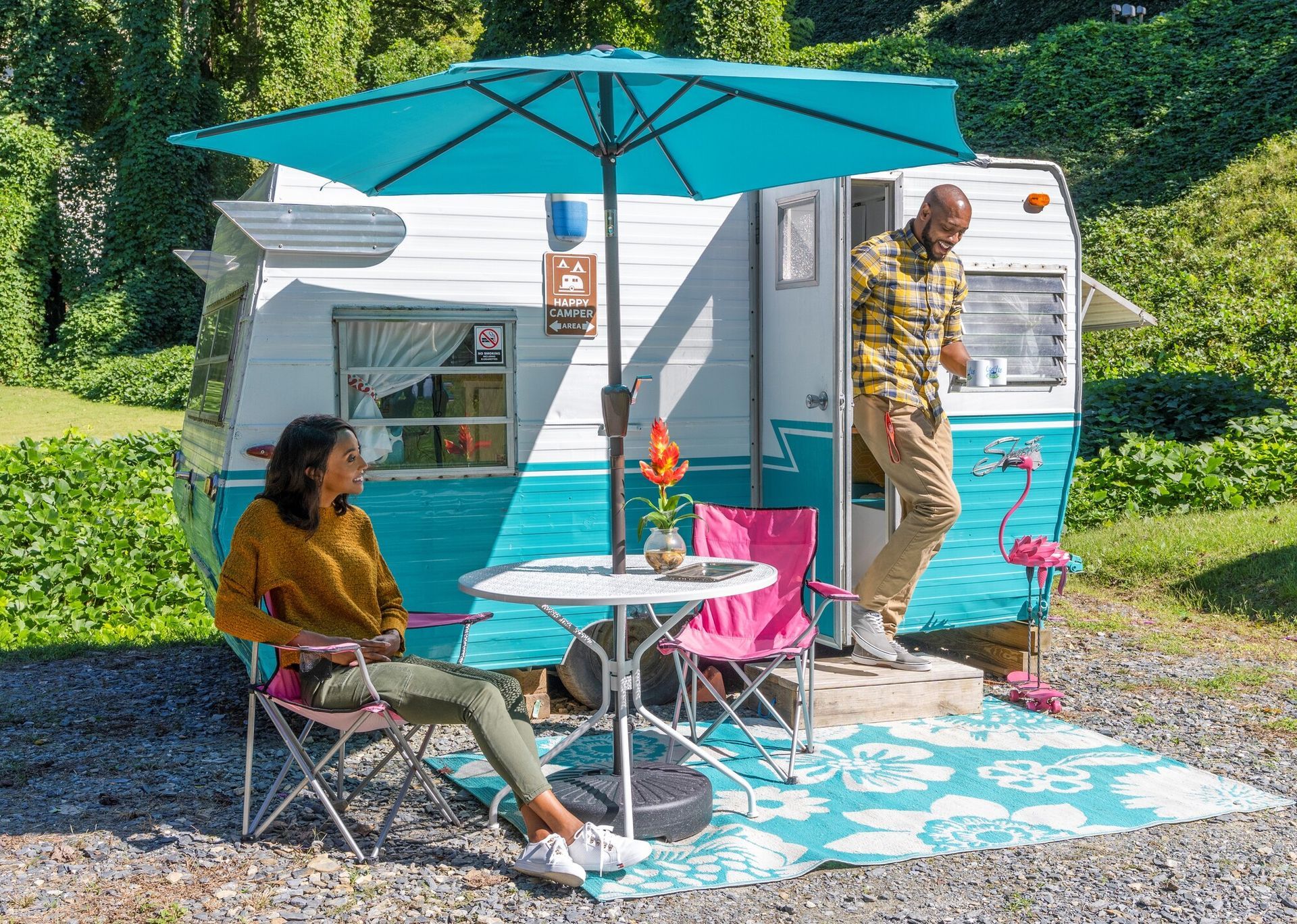Compact camper with turquoise umbrella; woman seated outside and another person stands in the doorway.